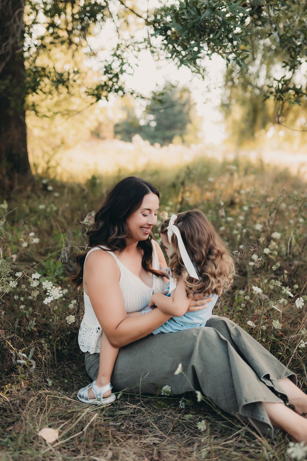A mother sits in a wildflower meadow holding her young daughter in warm golden sunlight.