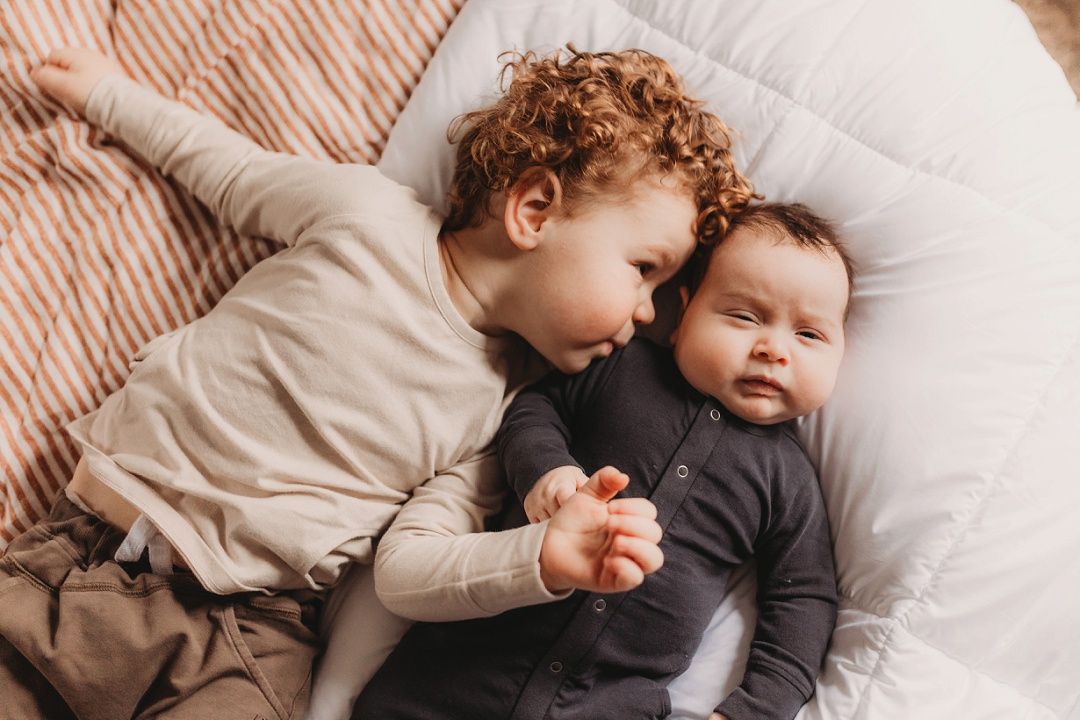 A curly-haired toddler gently kisses their younger baby sibling on a bed.