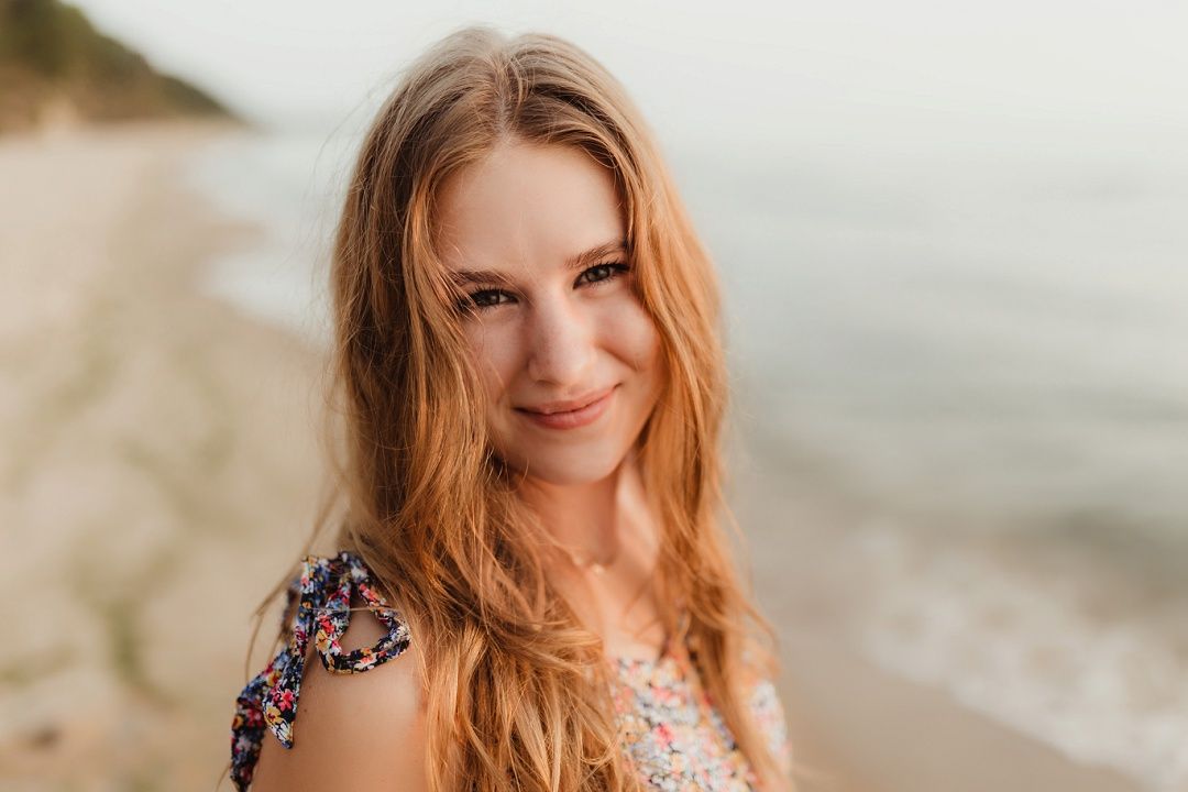 Young woman with long red hair smiling on beach at golden hour, ocean waves in background.