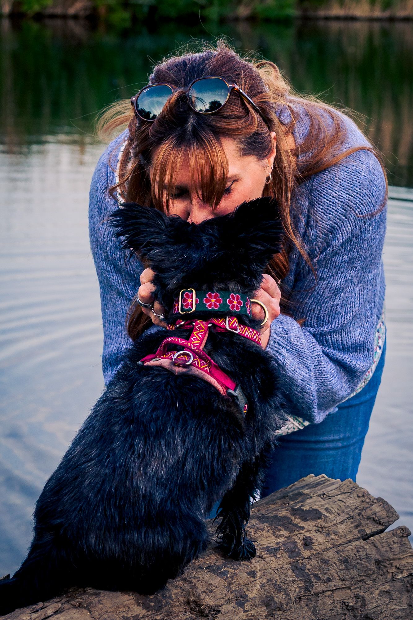 Woman in blue sweater hugging her black dog with pink collar near a serene lake.