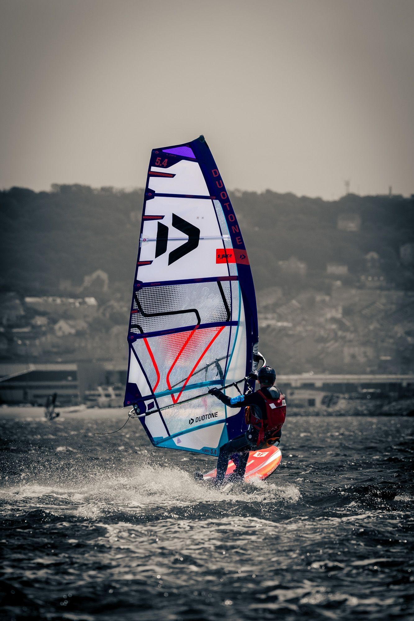A windsurfer rides choppy waves with a colorful Duotone sail against a grey coastal town backdrop.