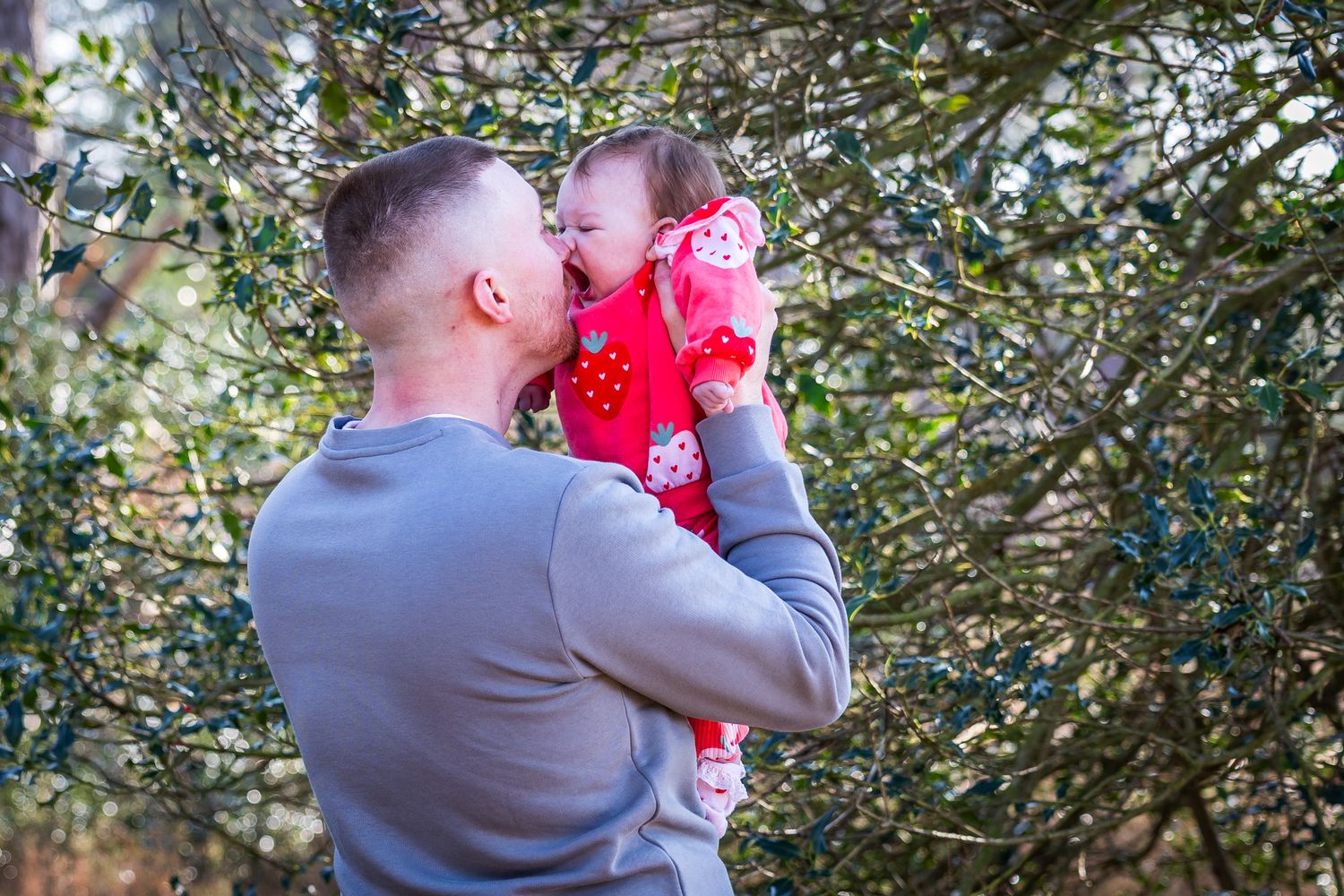 A man in a grey sweater lifts a baby girl in a red outfit among green trees, sharing a tender kiss in Dorset.
