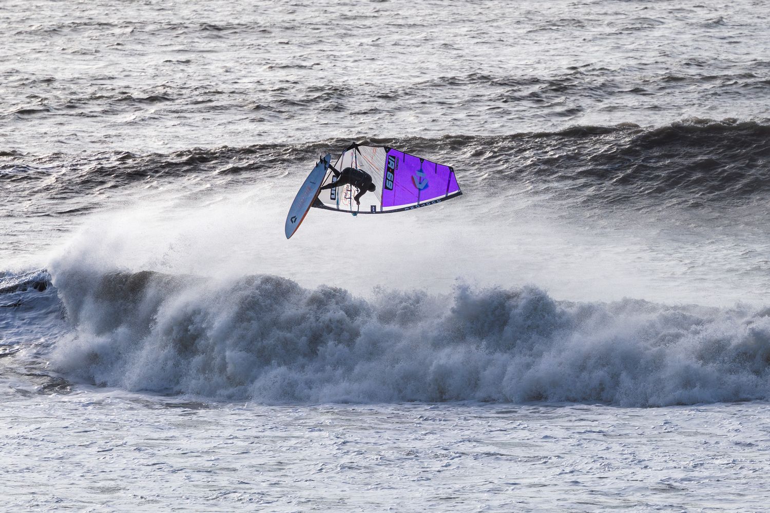 Windsurfer catching air above a crashing ocean wave with a purple sail during windy conditions at Kimmeridge, Dorset.