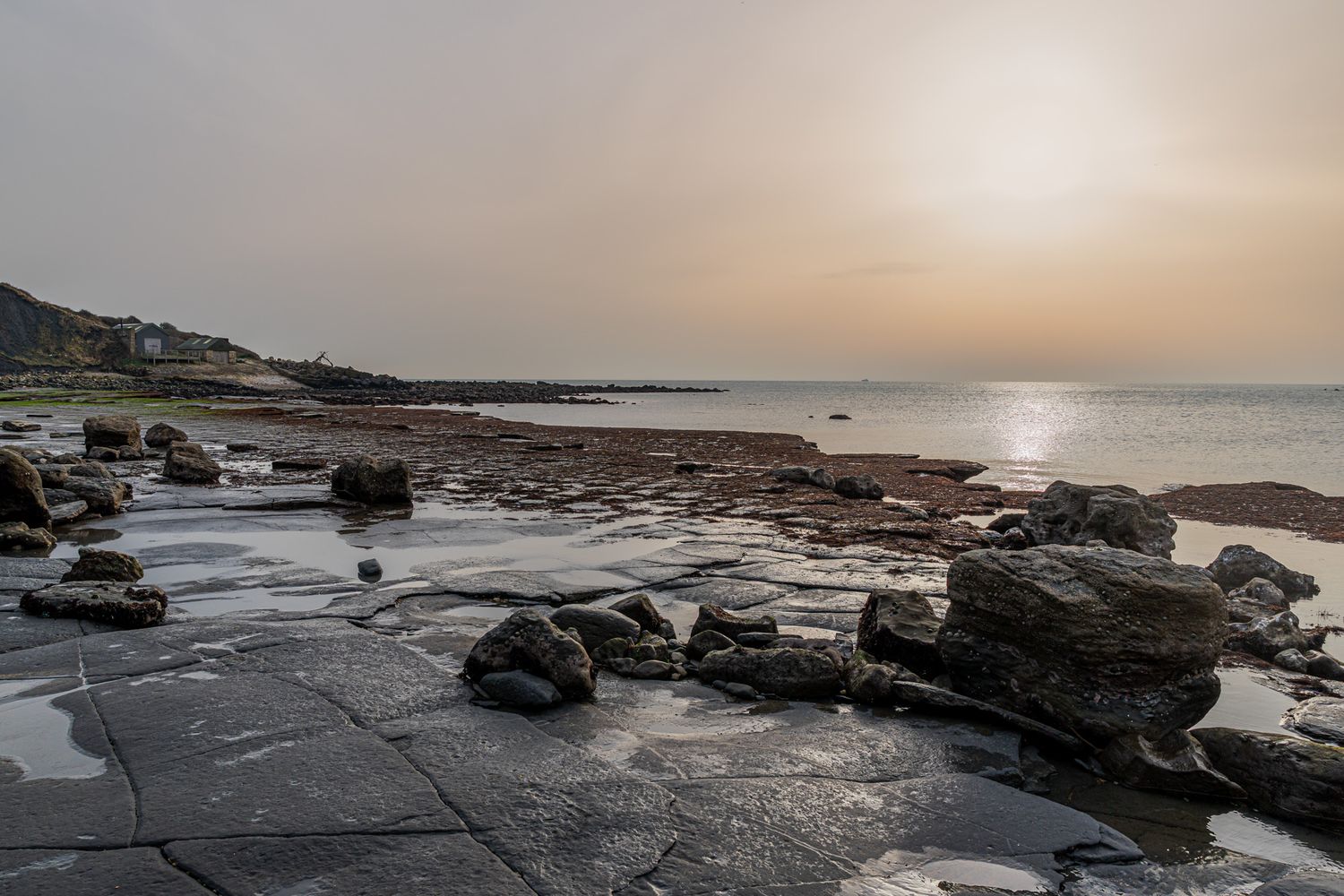 Rocky Dorset shoreline at sunset with flat stone slabs, scattered boulders, and calm sea reflecting golden light under a hazy sky.