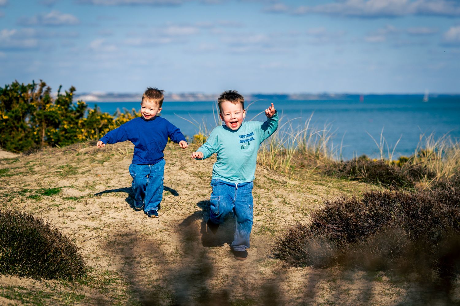 Two young boys joyfully running on a grassy coastal cliff with blue ocean and sky in the background at Studland Beach in Dorset.