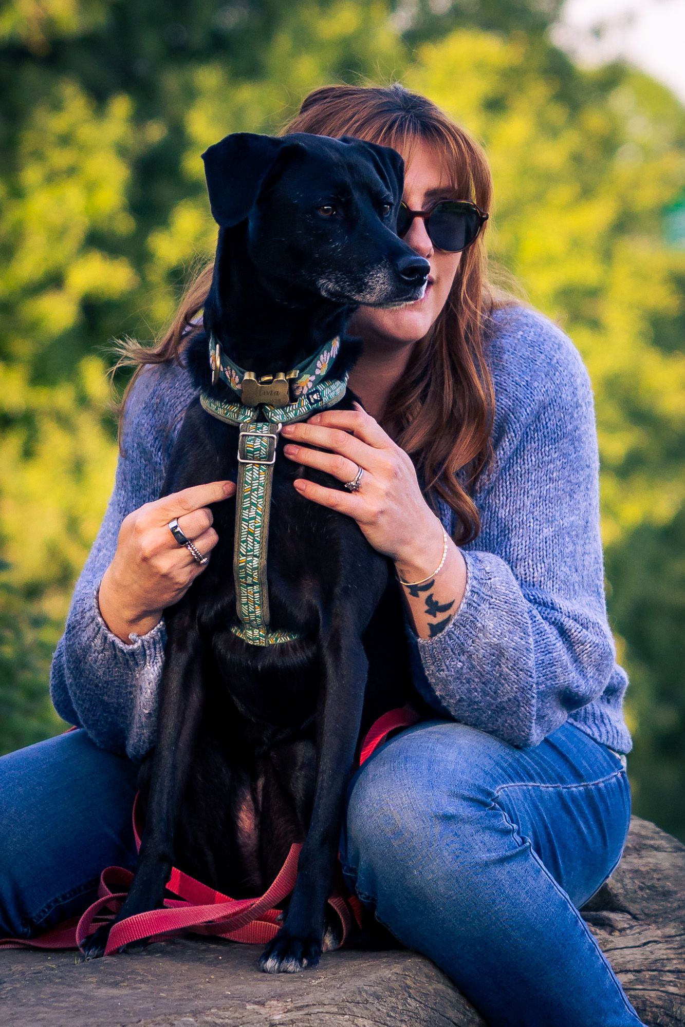 Woman in sunglasses hugging her black Labrador dog outdoors, both sitting on rocks with green trees in background.