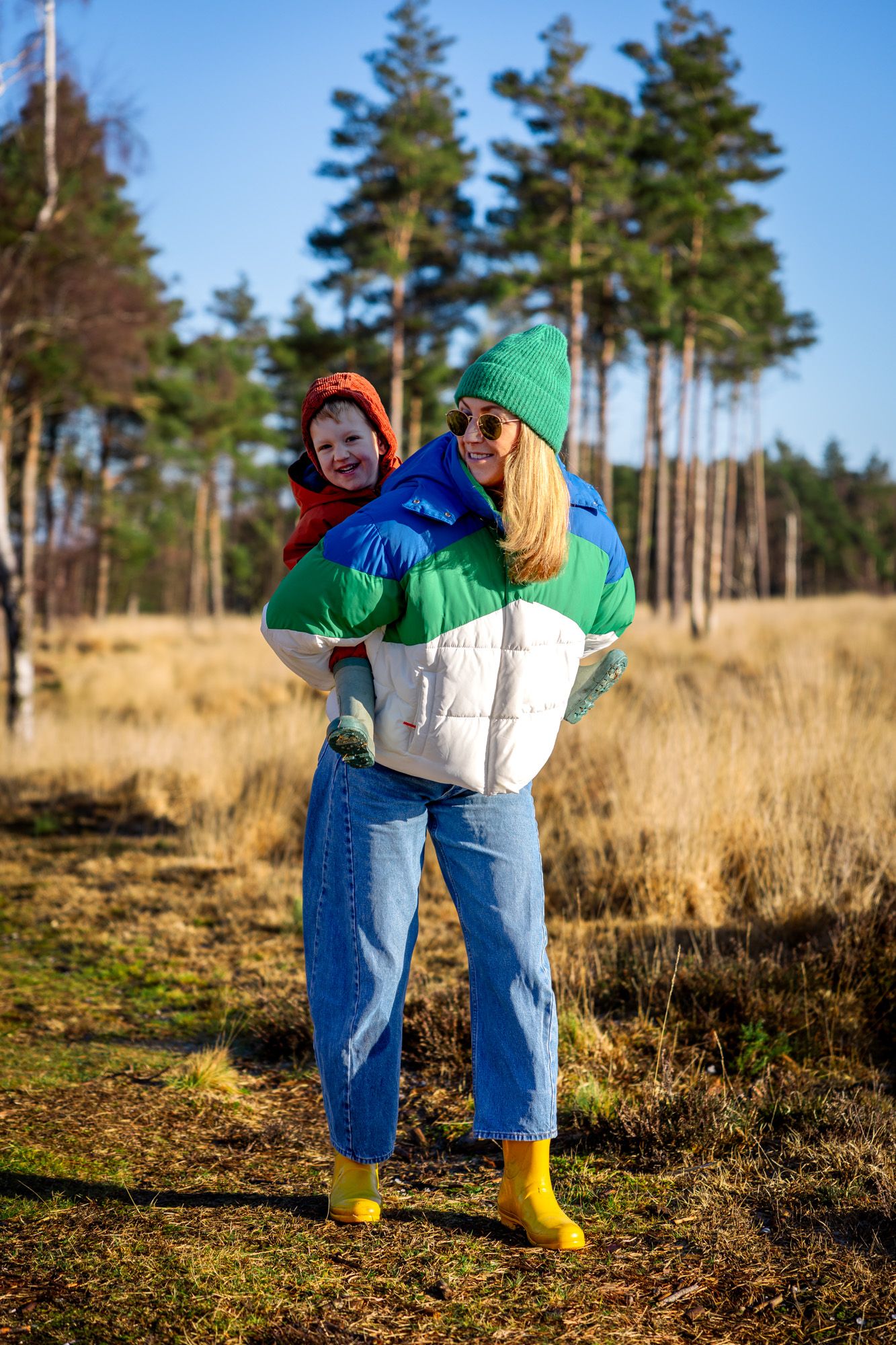 A woman in a colorful puffer jacket gives a child a piggyback ride outdoors near a pine forest in Dorset on a sunny day.