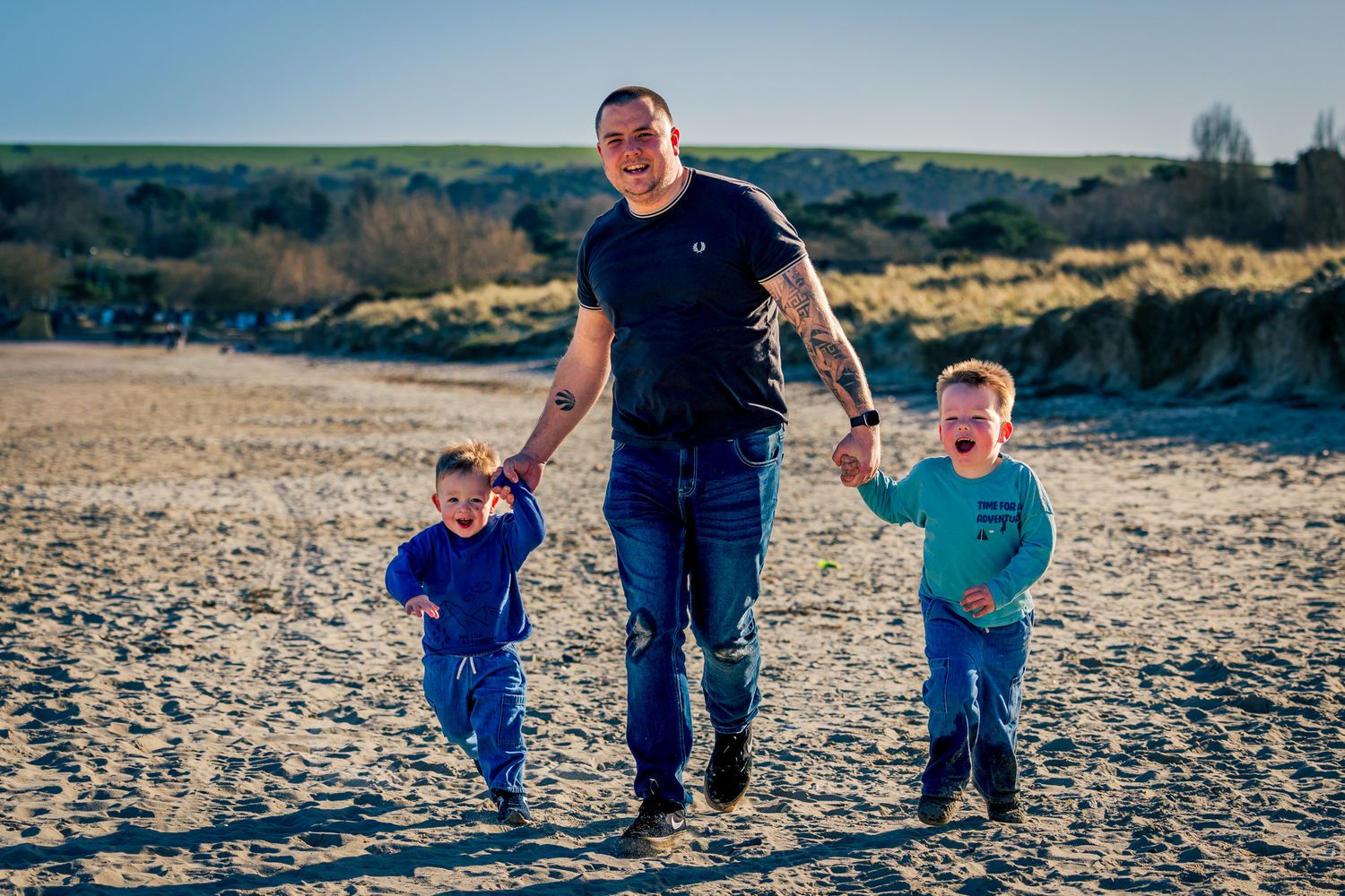 A father walks on a sandy beach in Dorset holding hands with his two young sons on a sunny day.
