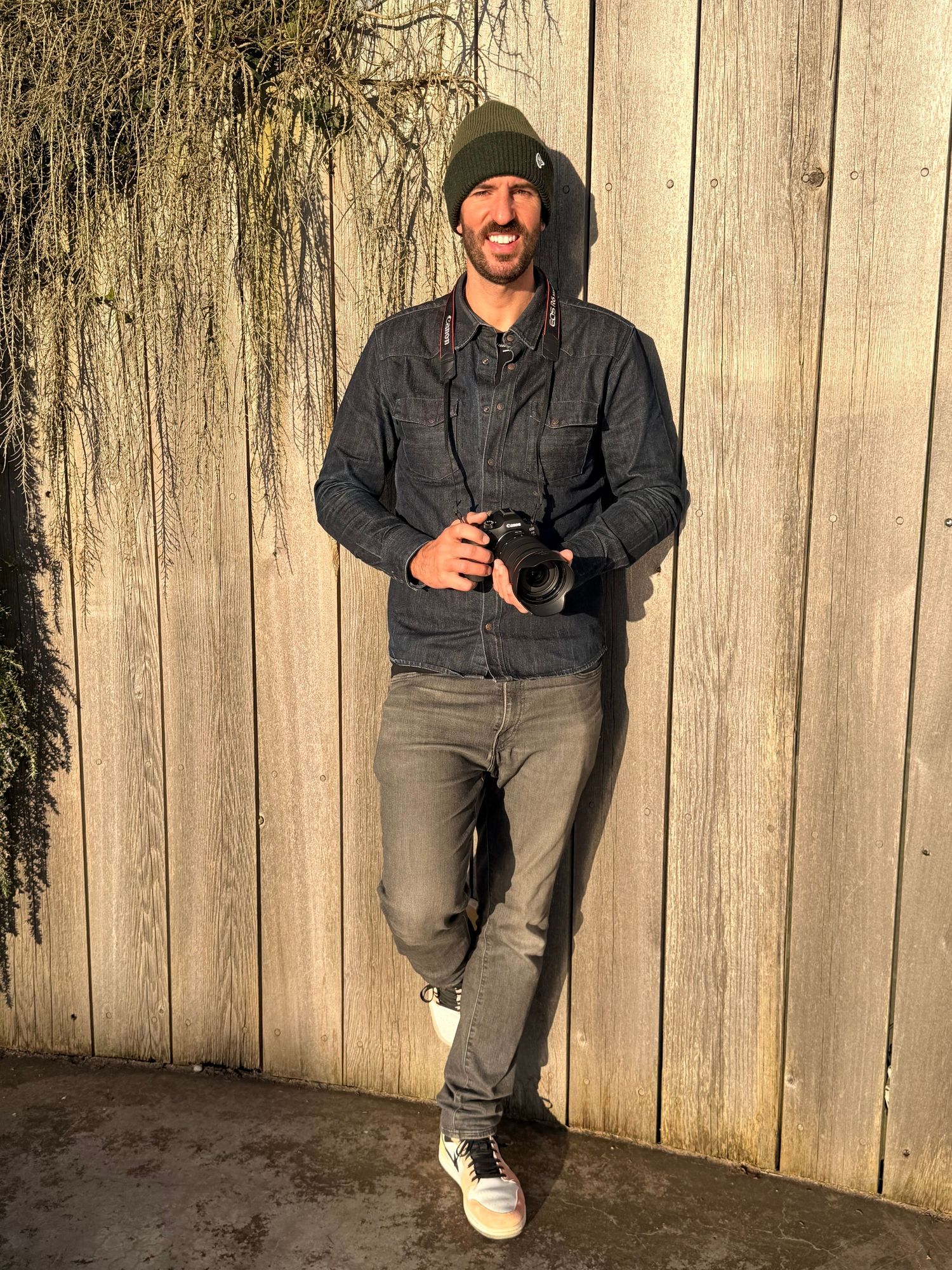 Philip J Long in a green beanie and denim jacket holding a camera, leaning against a wooden fence outdoors.
