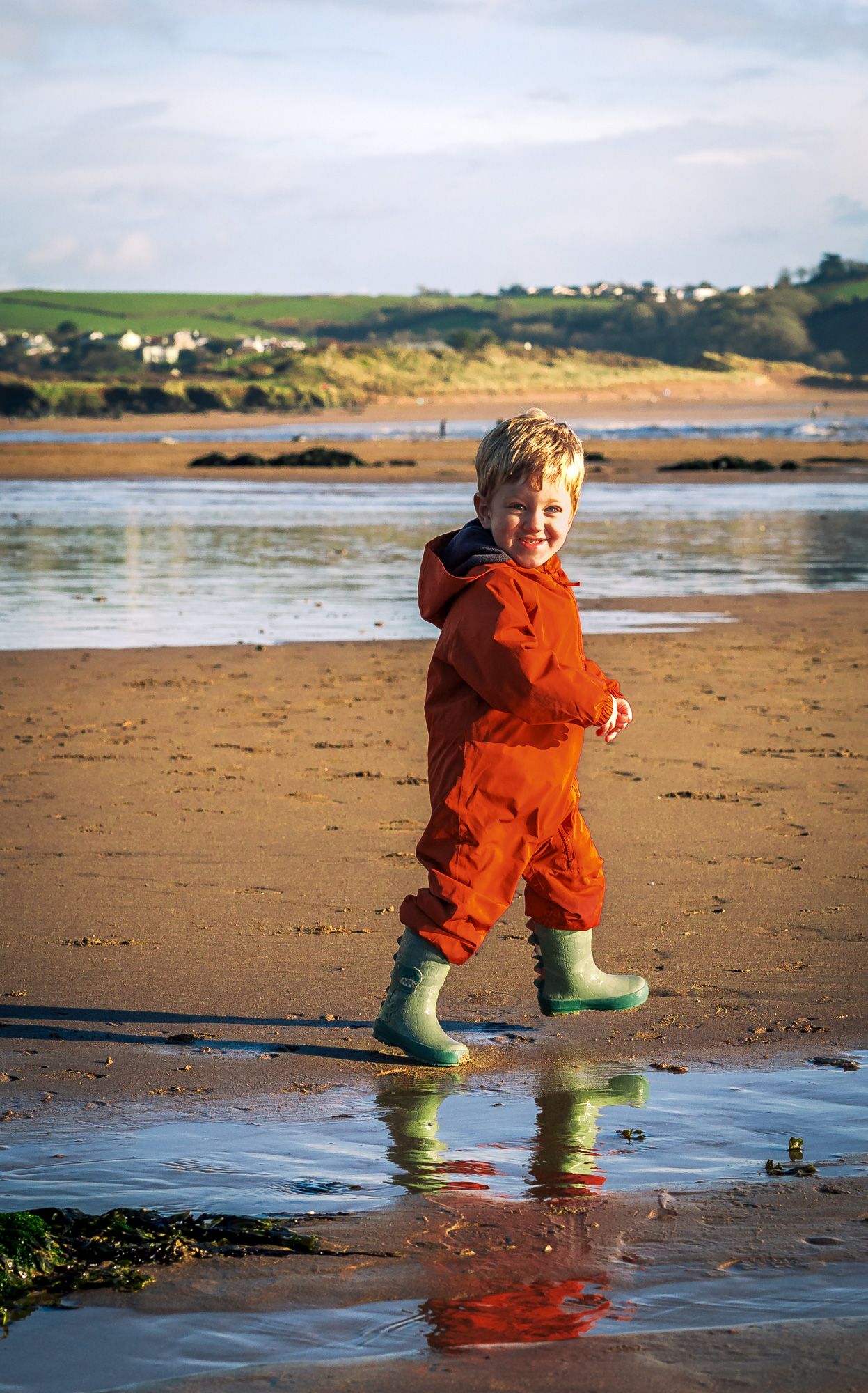 Happy young boy in red jumpsuit and green wellies splashing through puddles on a sandy beach.
