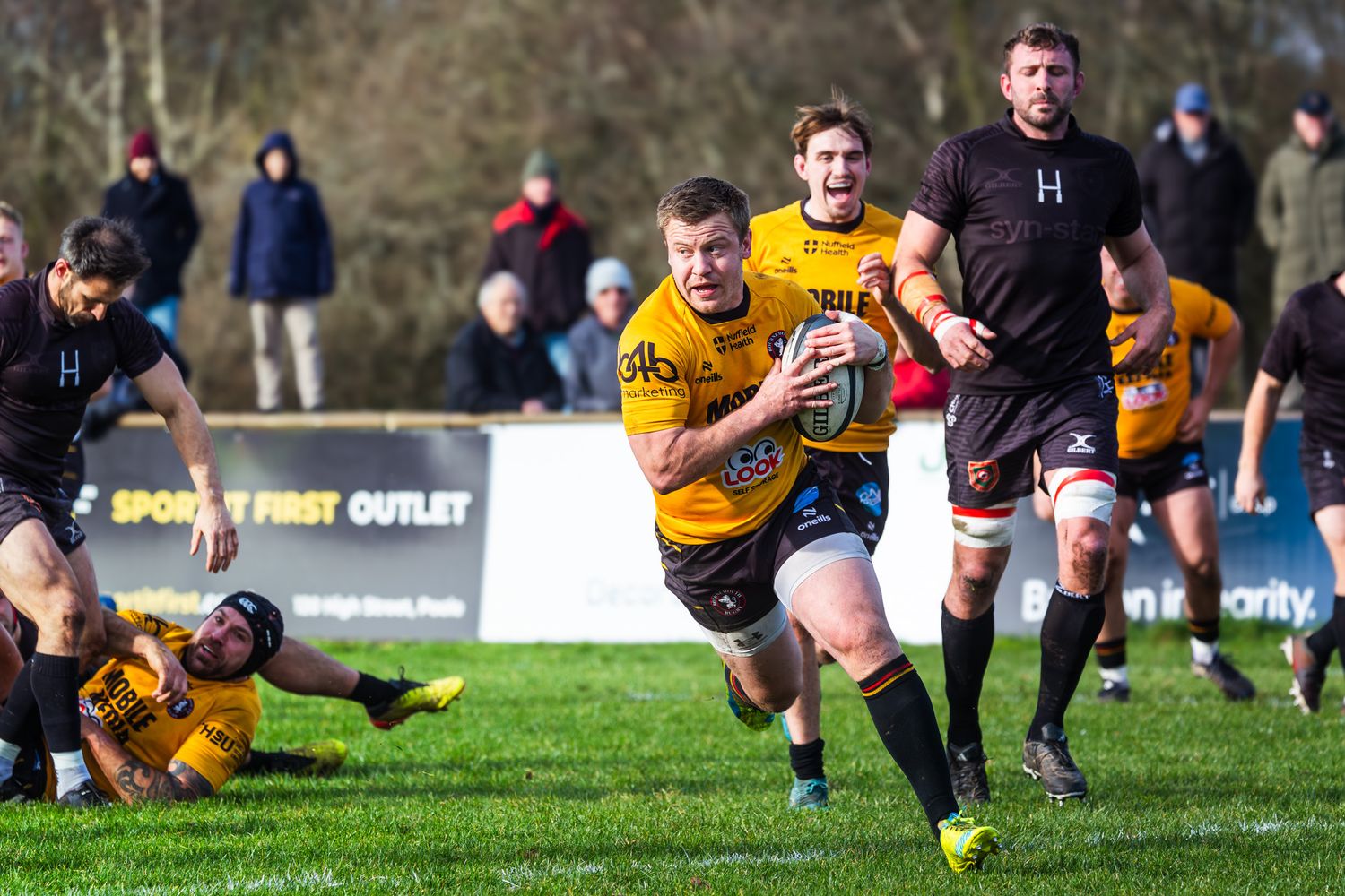 Rugby players compete in an outdoor match, with a player in yellow carrying the ball during a sunny afternoon game.