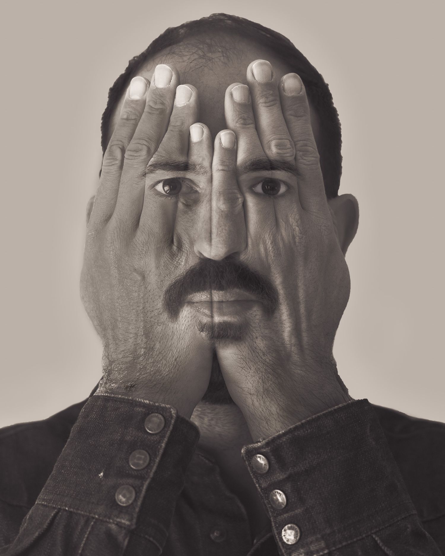 Black and white portrait of a man covering his face with both hands, revealing only his mustache and eyes.
