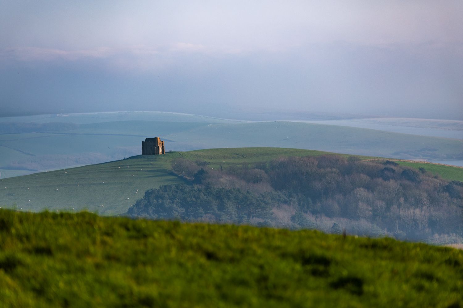 St Katherine's Chapel, Dorset, atop a lush green hill under a soft pastel sky with rolling countryside stretching into the distance.
