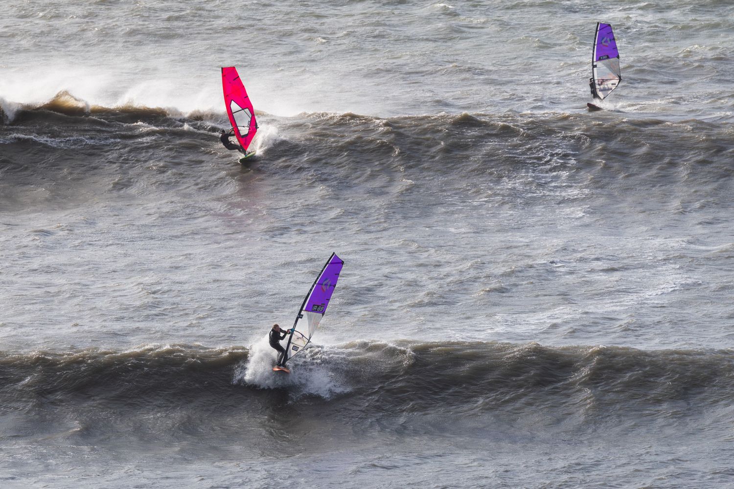 Three windsurfers ride large ocean waves with colourful sails in red, purple, and violet against choppy waters at Kimmeridge, Dorset.