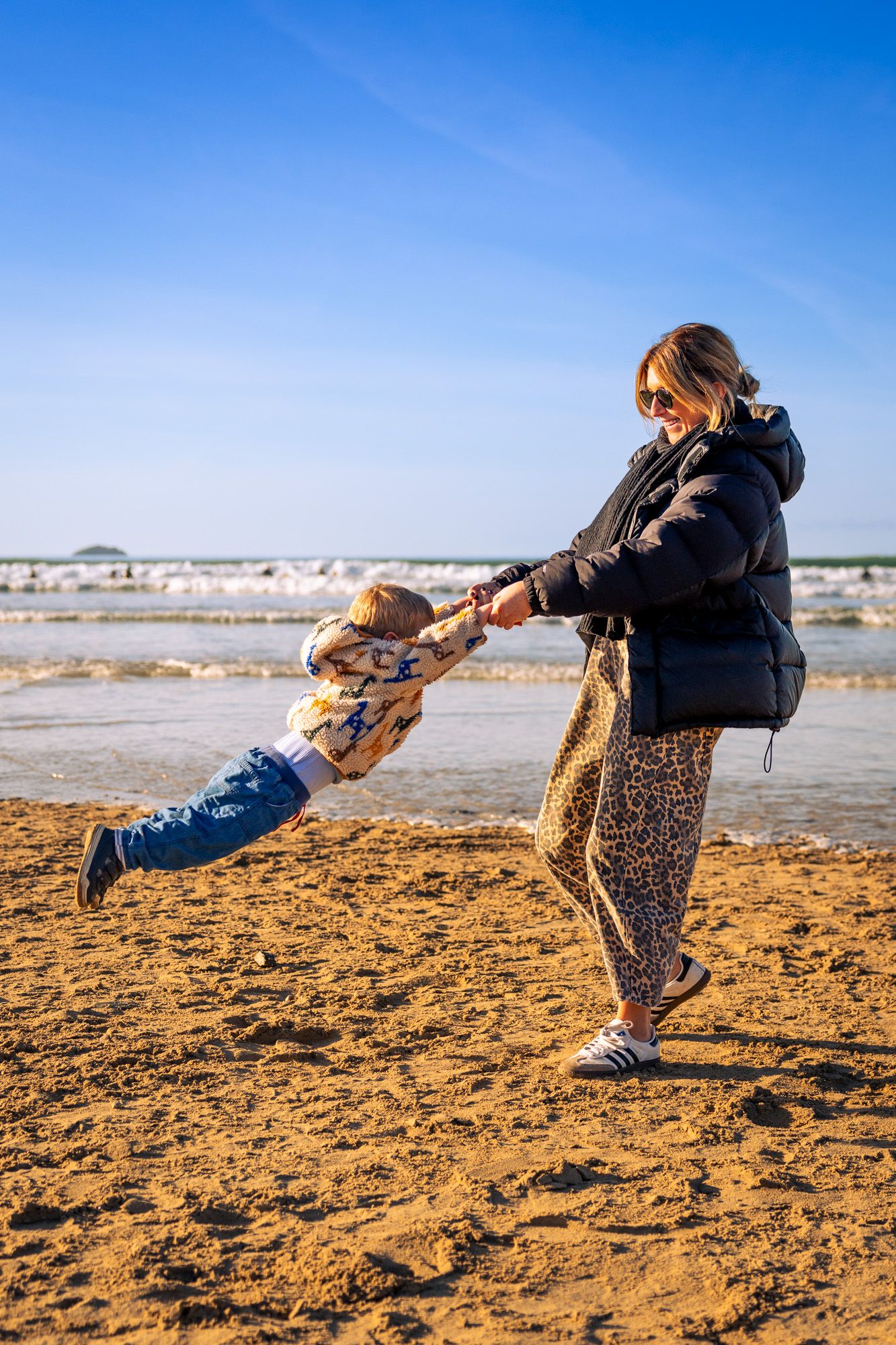 A woman in a black puffer jacket spins a young child by the hands on a sunny beach.