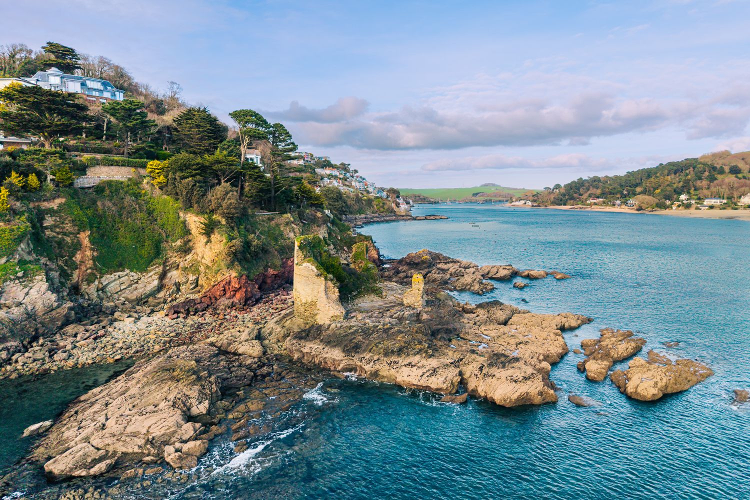 Aerial view of Salcombe Castle with turquoise waters, lush greenery, and charming seaside homes under a partly cloudy sky.