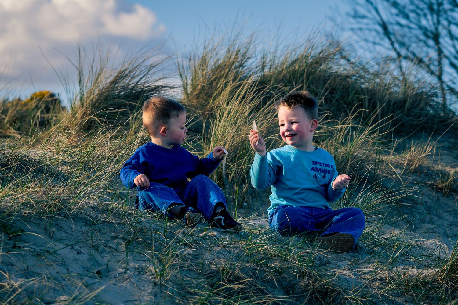 Two young toddler boys sitting and laughing together on sandy grass dunes in Dorset under a blue sky.