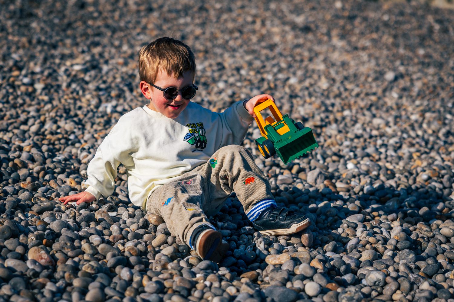 A young boy wearing sunglasses sits on a pebble beach playing with a colorful toy truck outdoors.