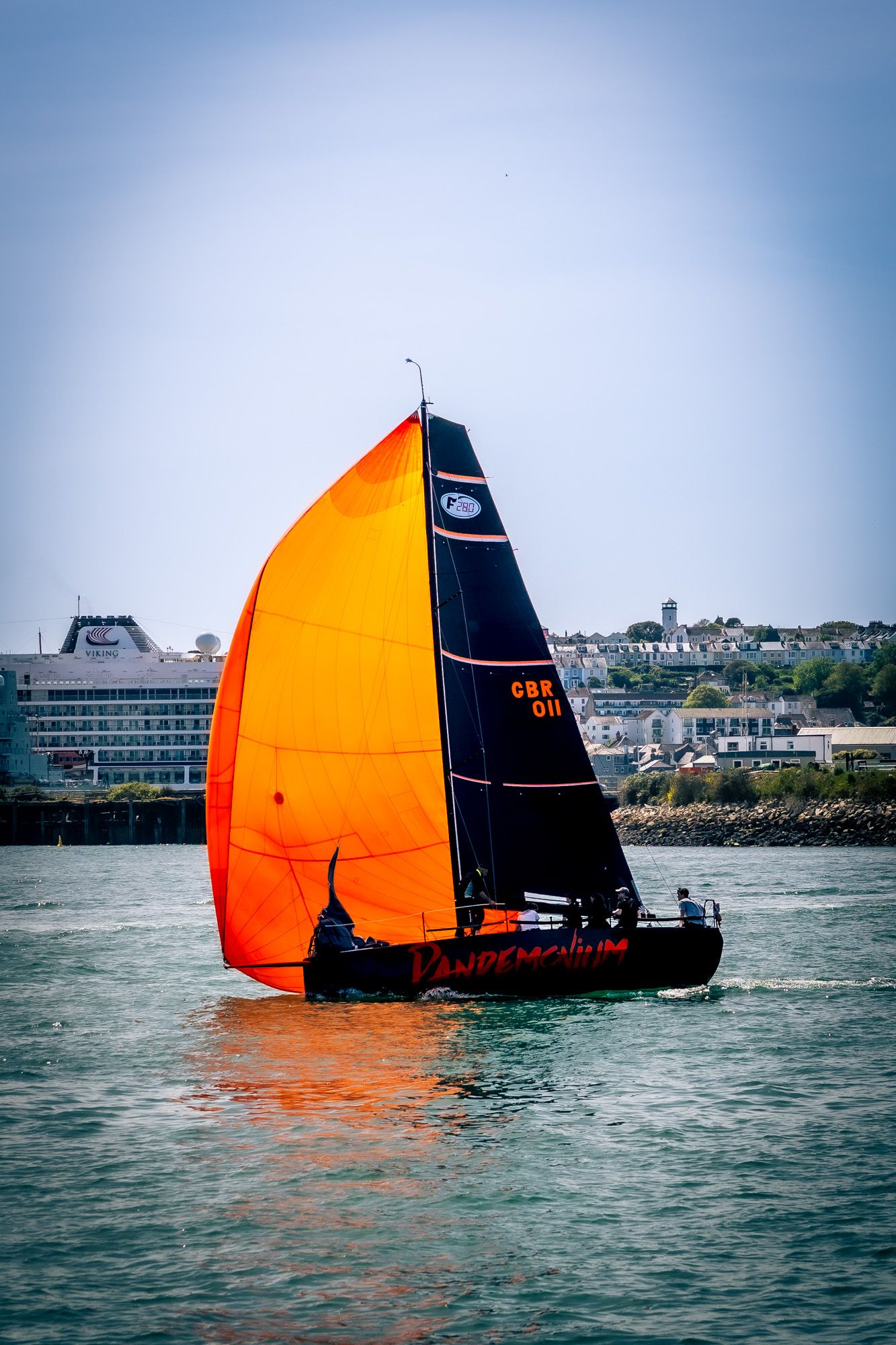 Black sailboat with vibrant orange spinnaker sail racing on blue water with coastal town and cruise ship in background.