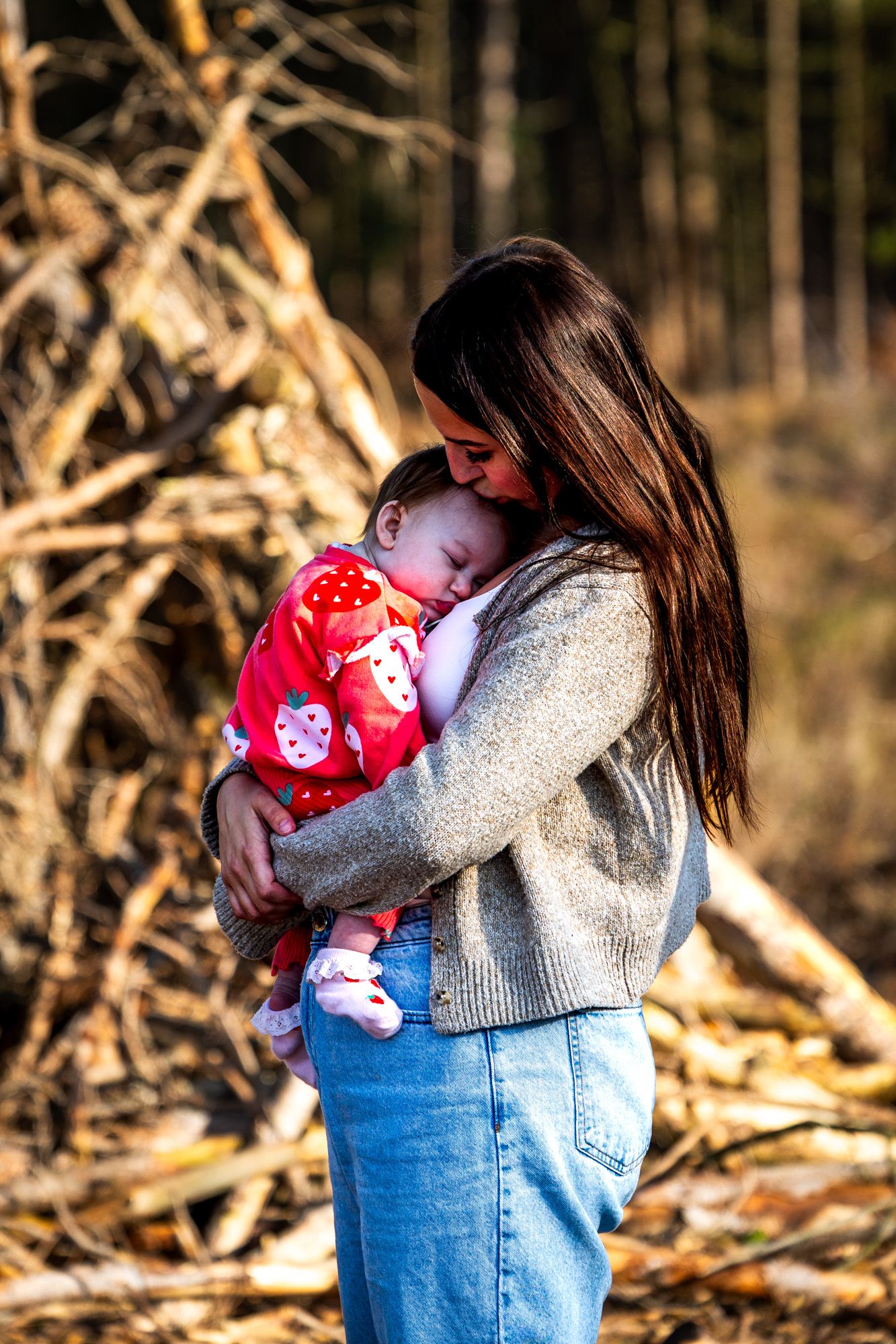 Young mother tenderly kissing sleeping baby dressed in red outdoors in autumn forest setting in Dorset..
