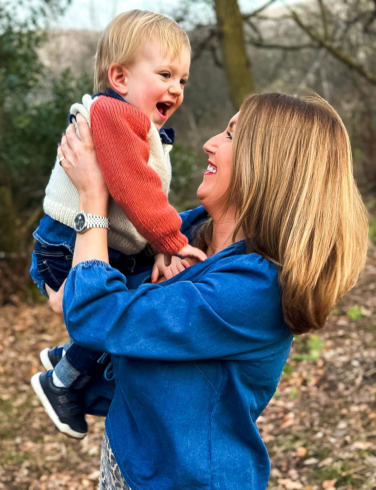 A joyful woman in a blue jacket lifts a laughing toddler in a red sweater outdoors in Dorset.