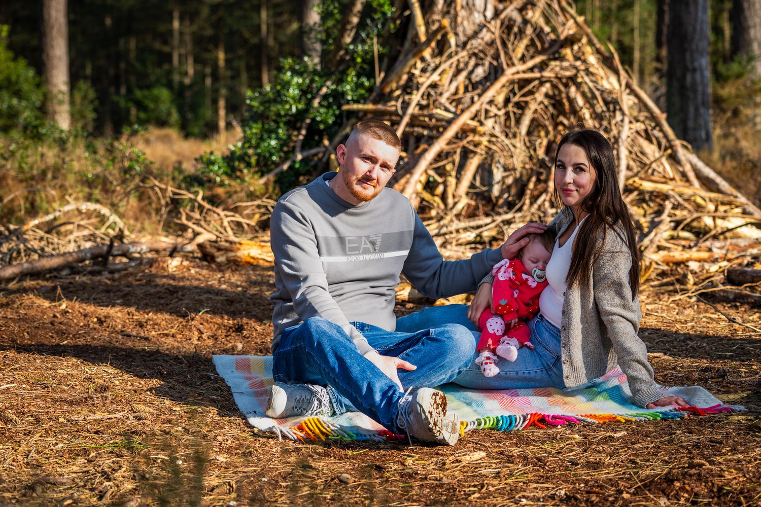 A couple sits on a colorful blanket in a forest, holding a baby, with a rustic log structure behind them in Dorset.