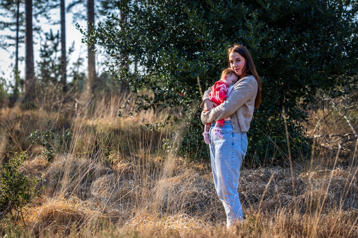 Woman holding a baby in a red outfit standing in a natural field in Dorset with pine trees in golden sunlight.