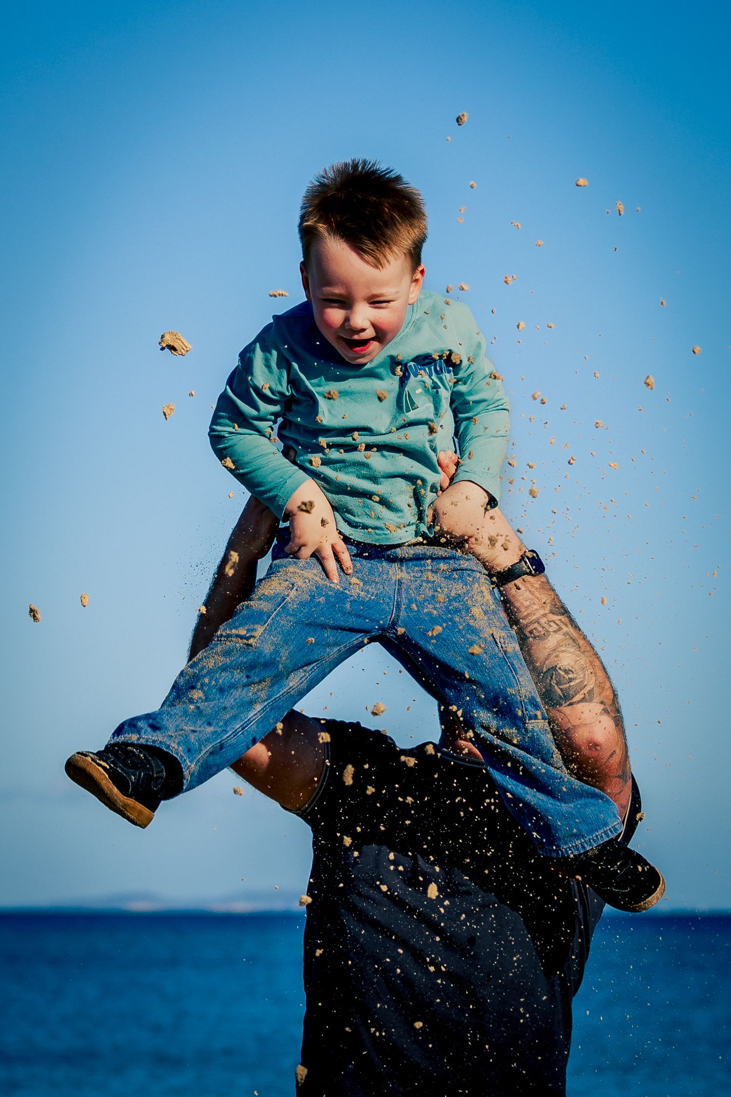 Laughing child being lifted high in the air by adult on sandy beach under blue sky.
