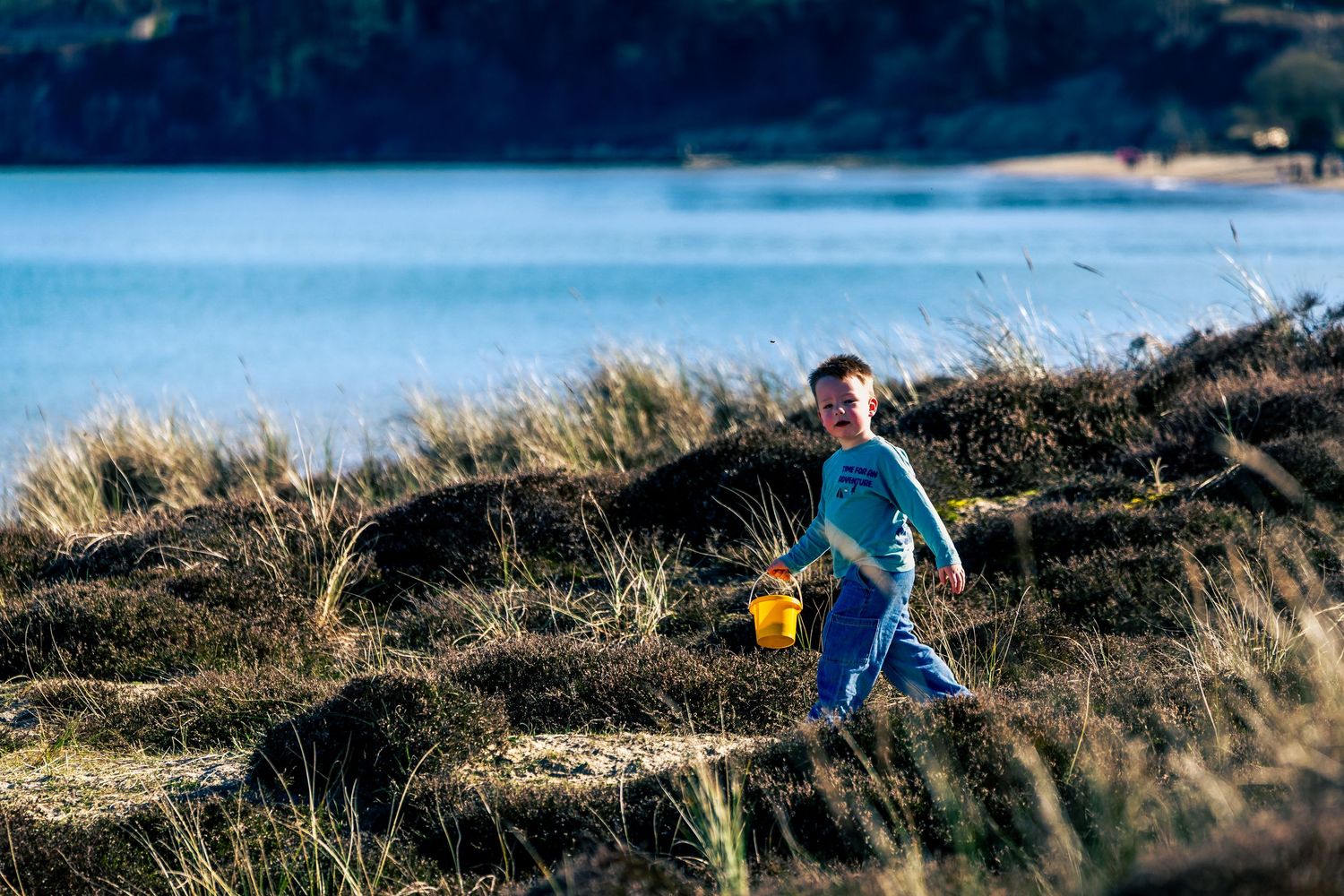 Young boy carrying yellow bucket walking through coastal grassland beside the sea in Dorset.
