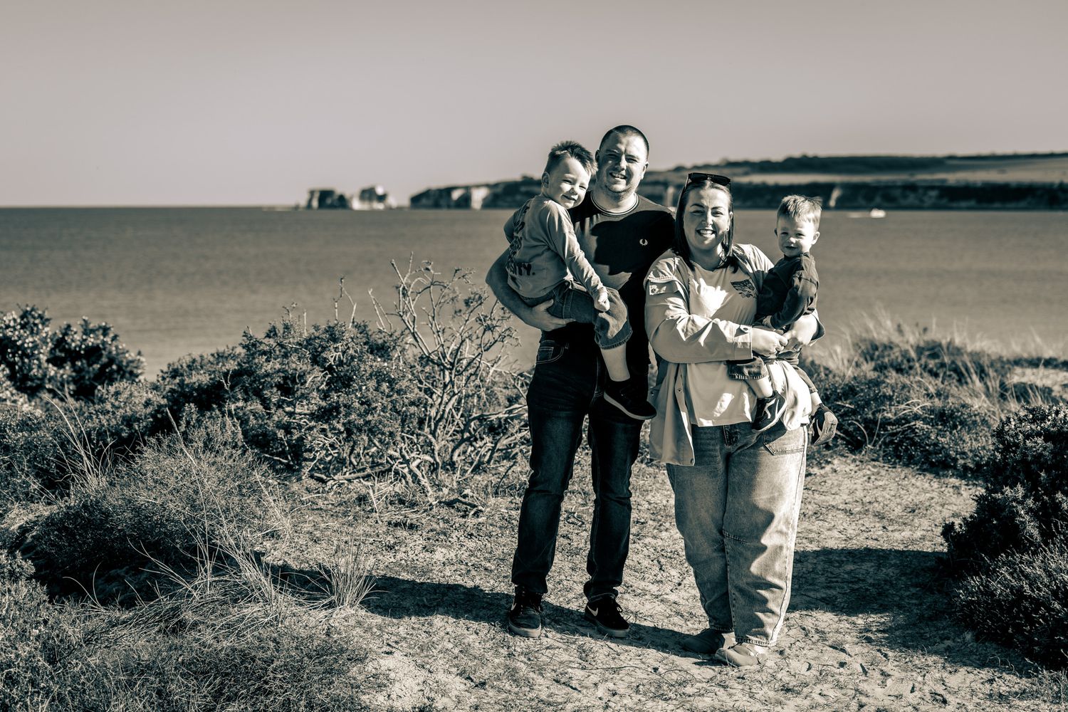 Black and white photo of two adults holding children on a coastal cliff path in Dorset with sea and cliffs in the background.