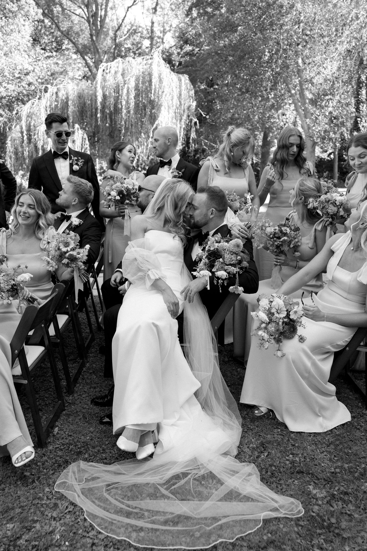 Black and white photo of bride and groom sharing romantic kiss surrounded by wedding party outdoors.