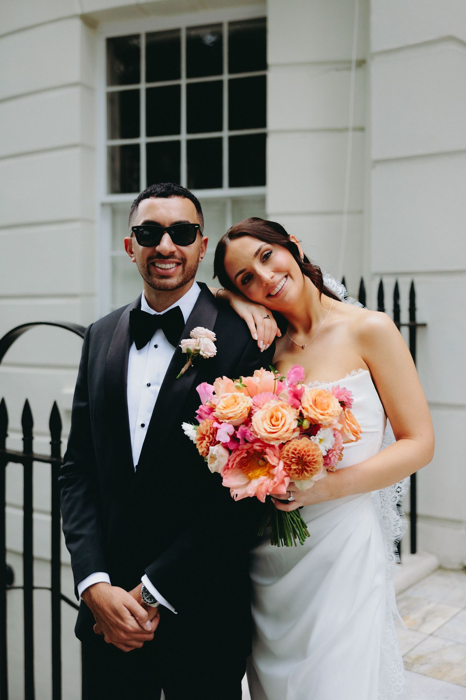Smiling couple in wedding attire pose outside white building with vibrant pink and peach bouquet.