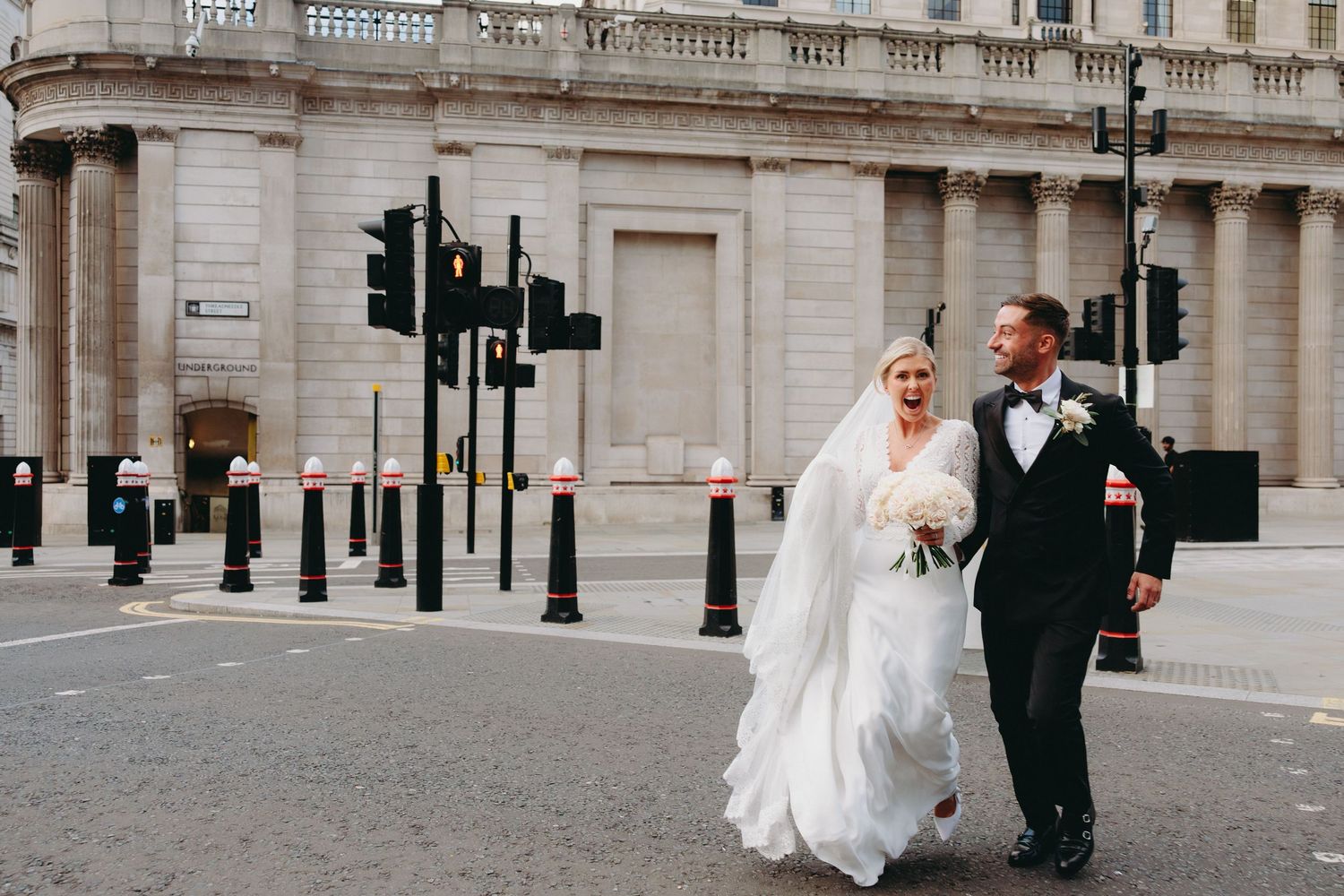 Bride in white gown and groom in black tuxedo walk hand in hand across urban city street.