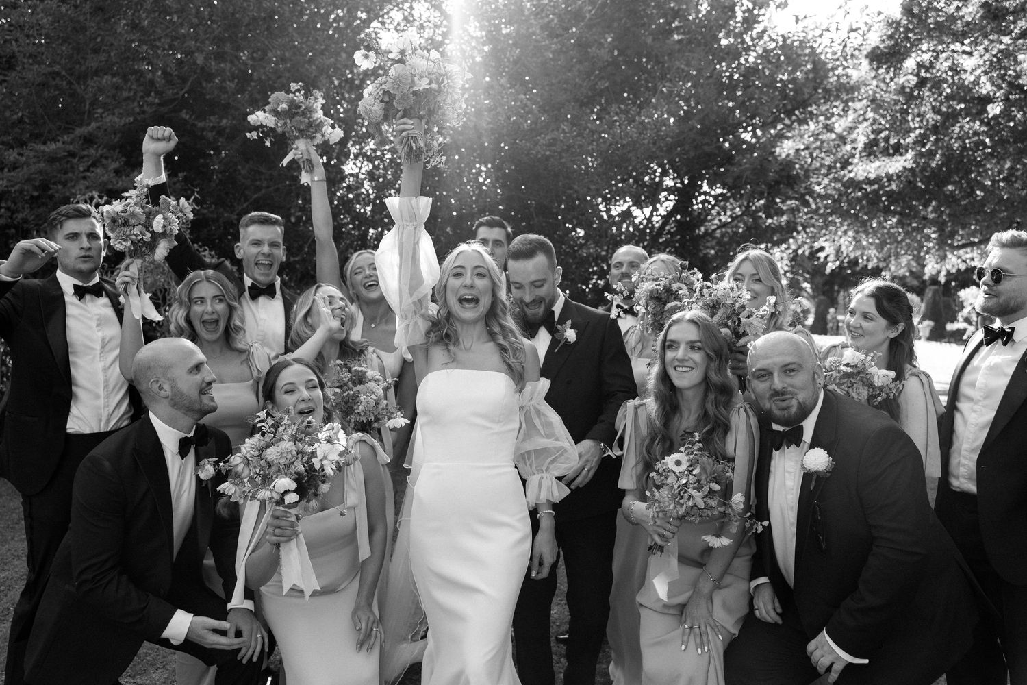 Bride and groom celebrate joyfully with wedding party in black and white outdoor photo.