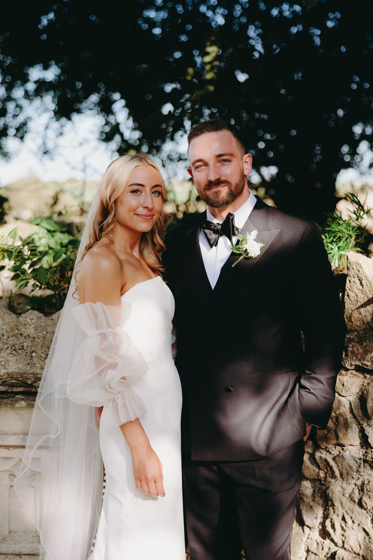 Blonde bride in off-shoulder gown with tulle sleeves poses with bearded groom outdoors.
