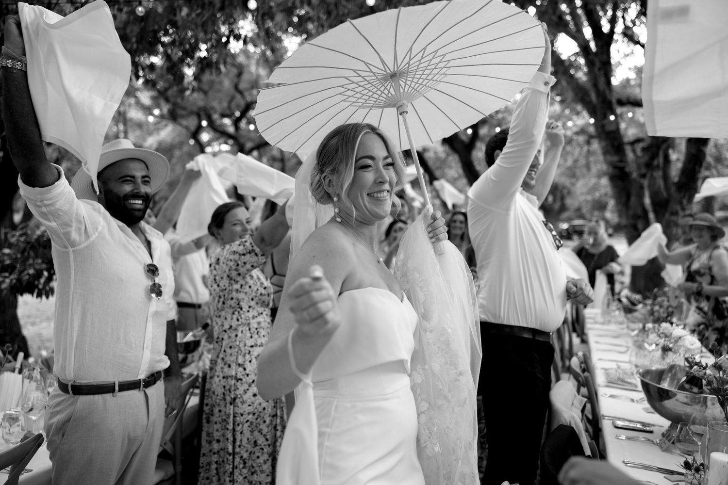 Smiling bride holds white parasol surrounded by cheering guests at outdoor wedding reception.