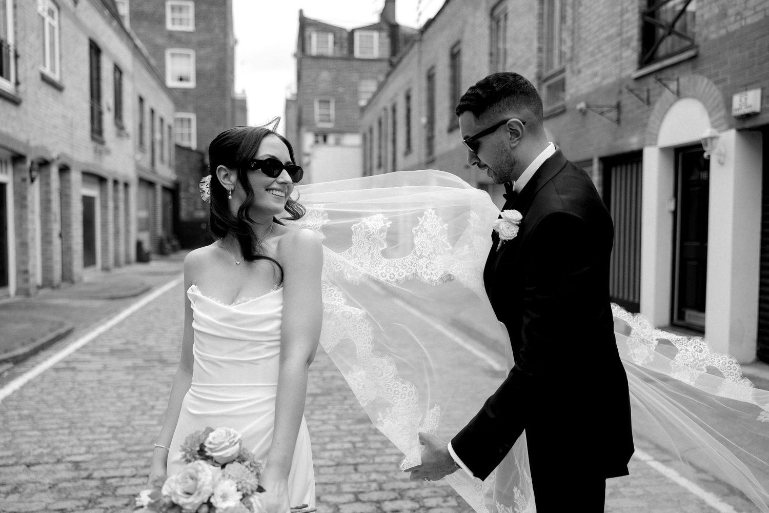 Bride with veil blowing in wind stands beside groom on cobblestone street in black and white.