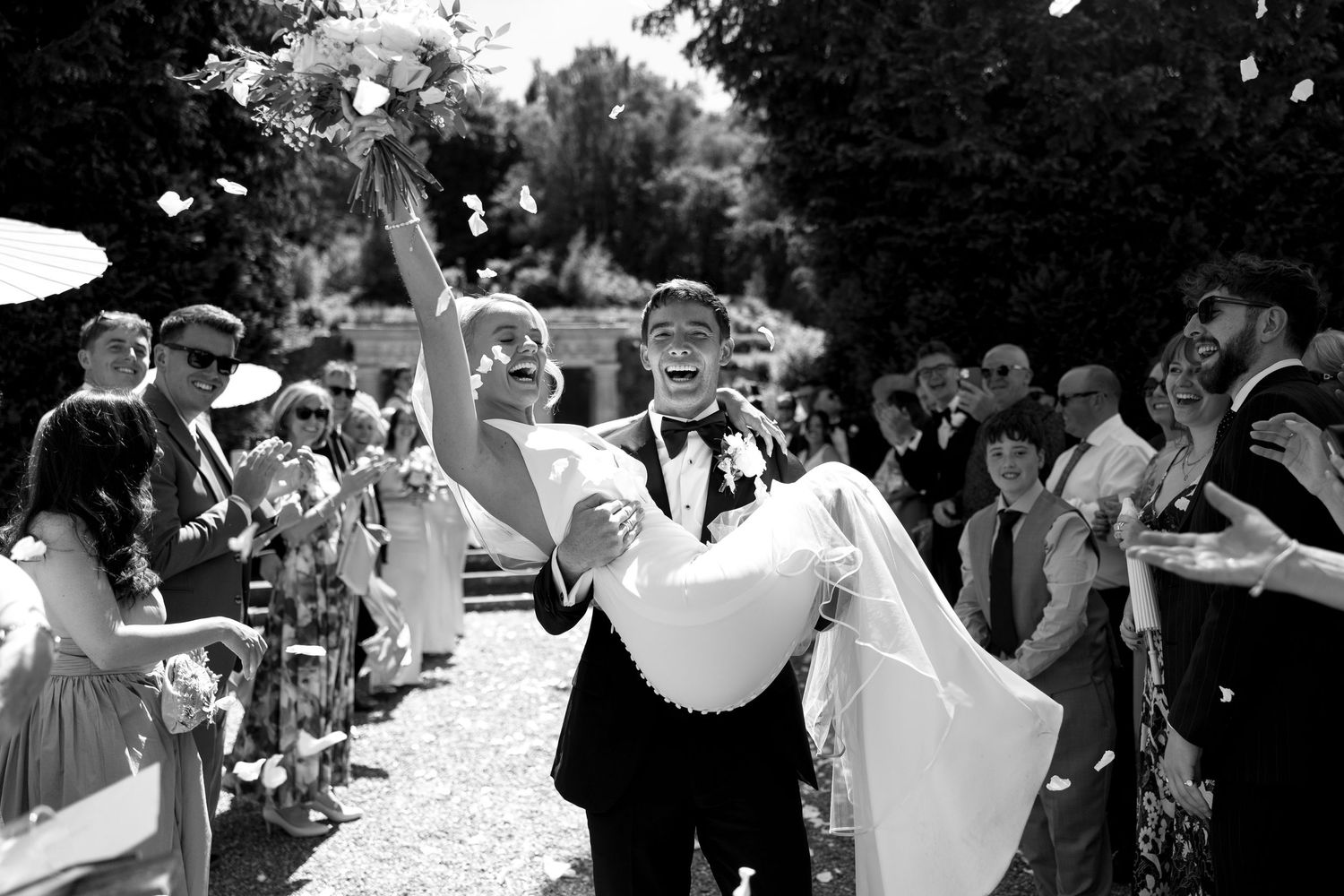 Groom lifts bride while guests celebrate with confetti at outdoor wedding ceremony.