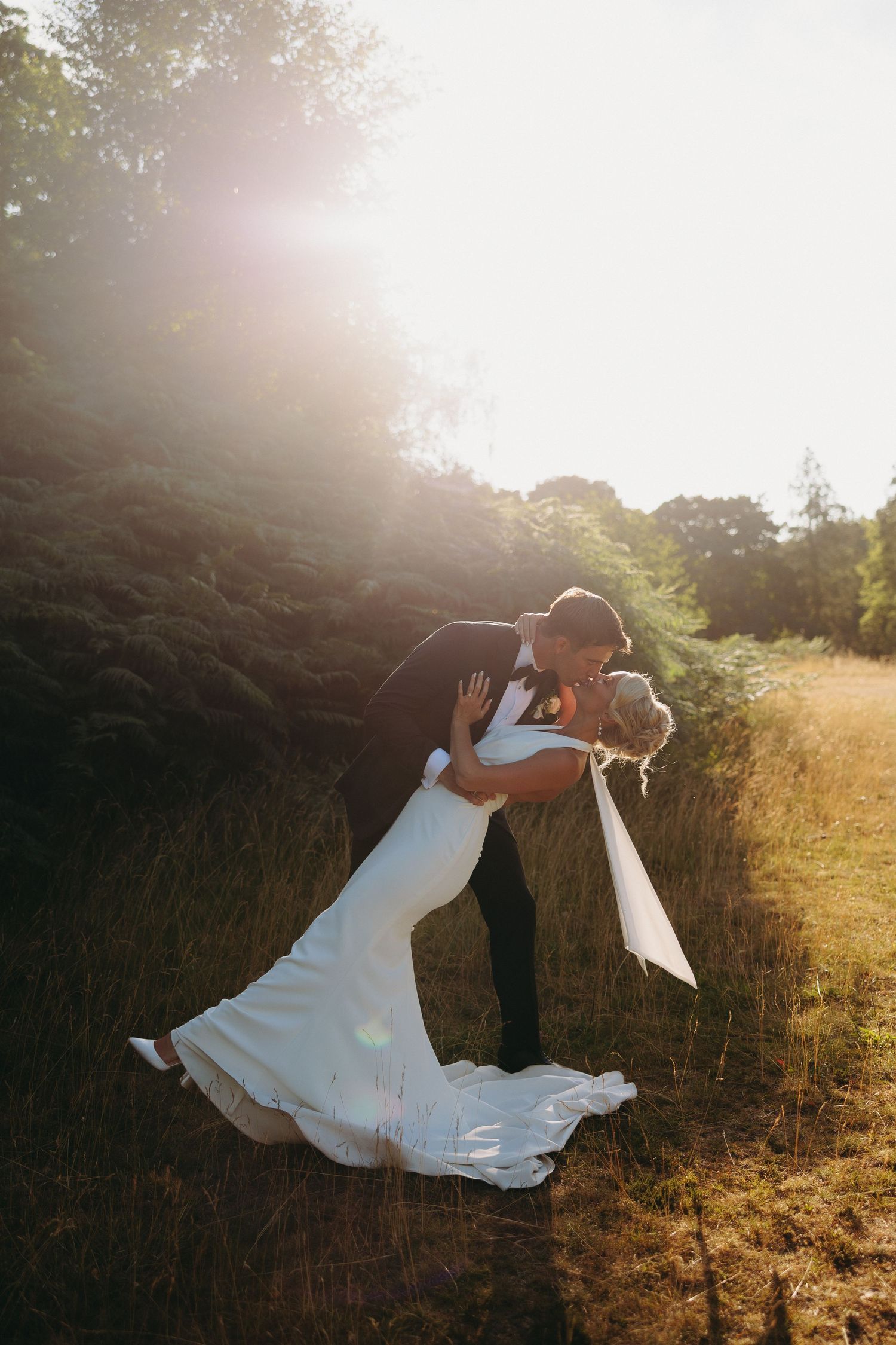 Groom dips and kisses bride in white mermaid gown in golden sunlit field at romantic wedding.
