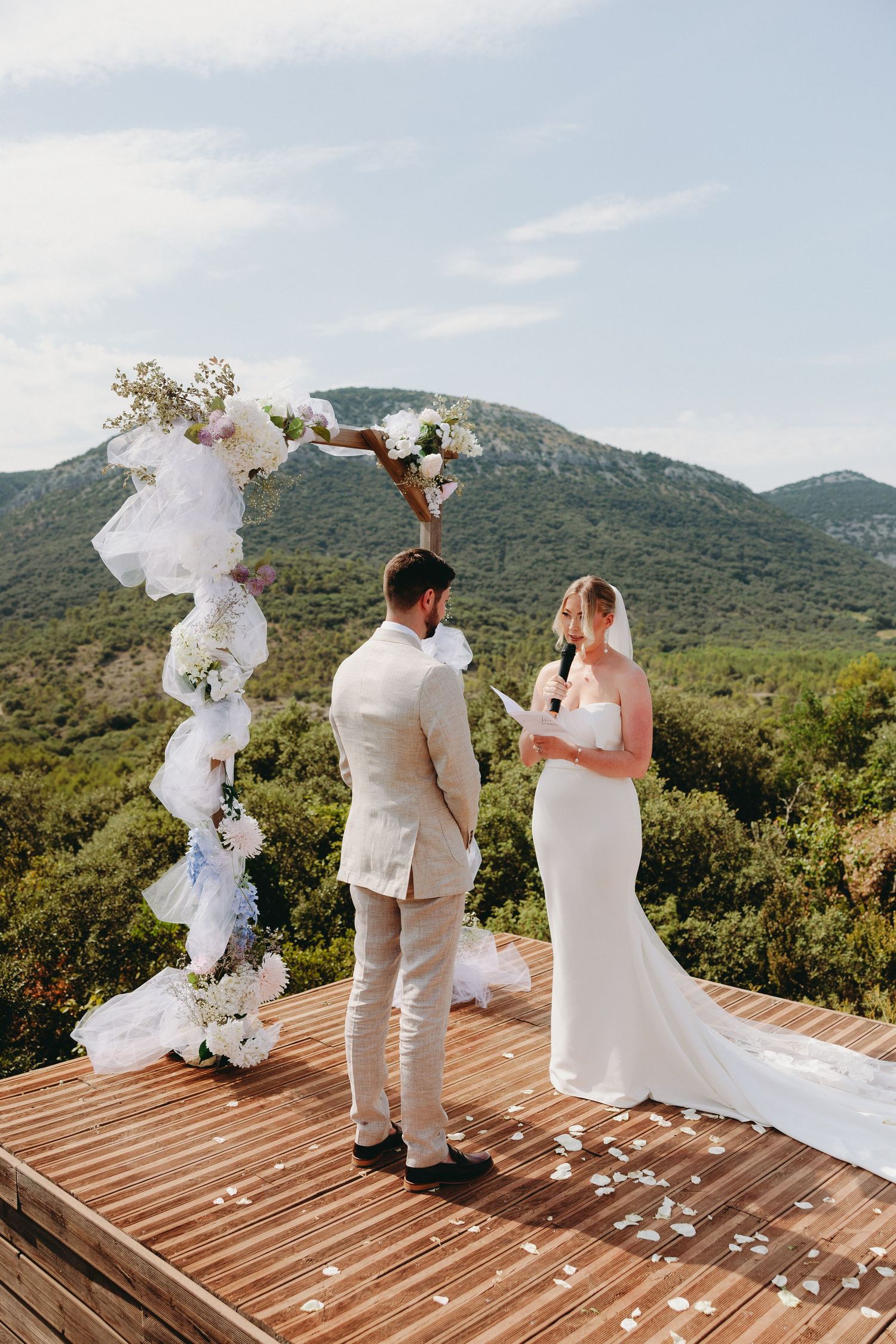 Bride reads vows to groom at outdoor ceremony with floral arch and mountain backdrop.