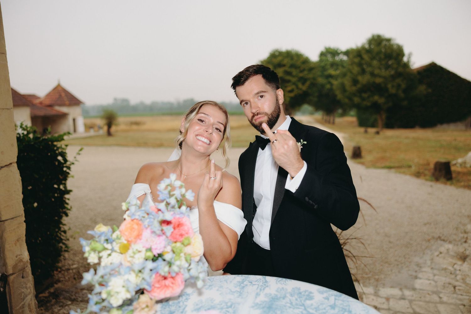 Smiling bride and groom show off wedding rings while posing outdoors at their countryside wedding venue.