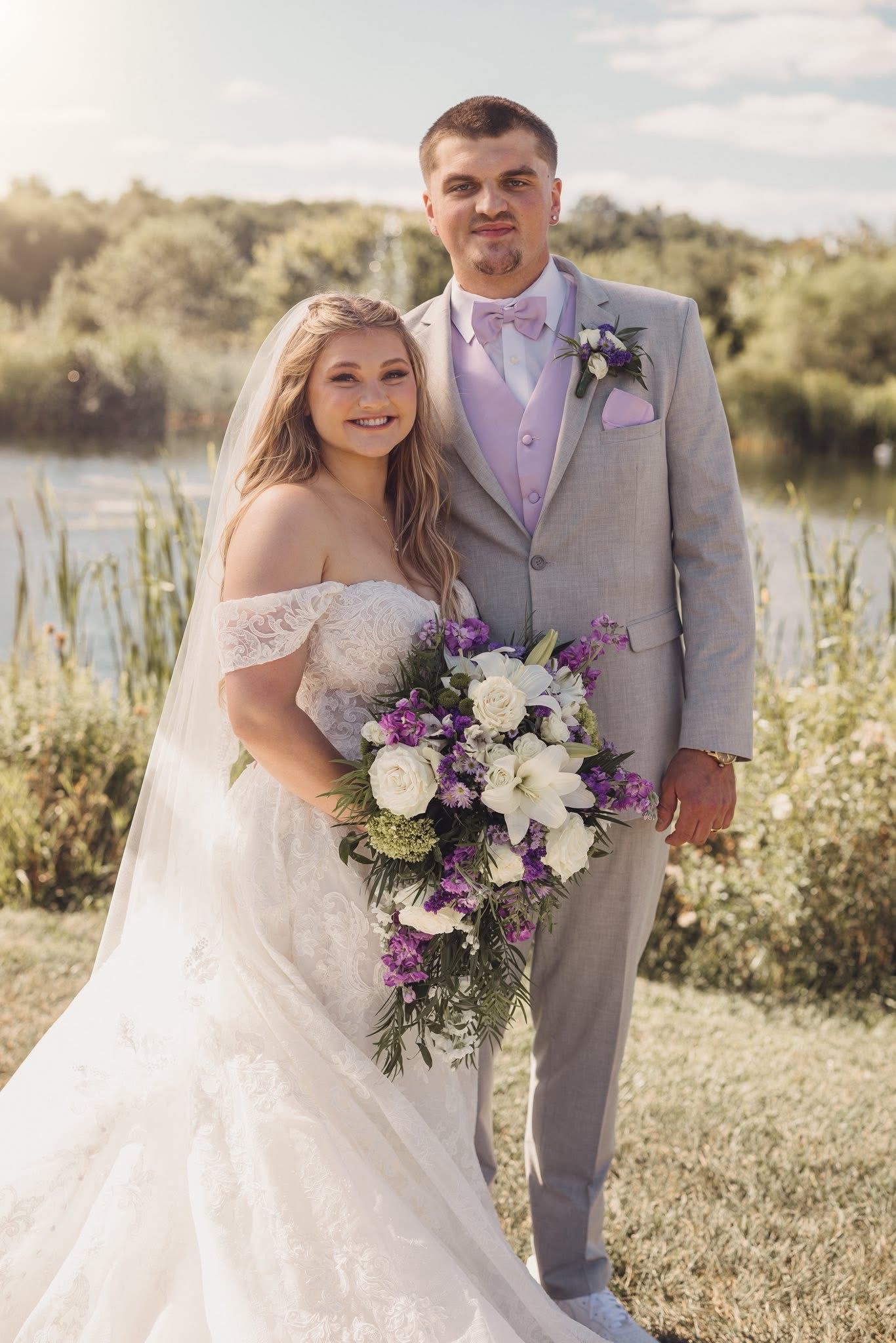 Wedding portraits in a sunlit meadow with purple and white floral bouquet accents.