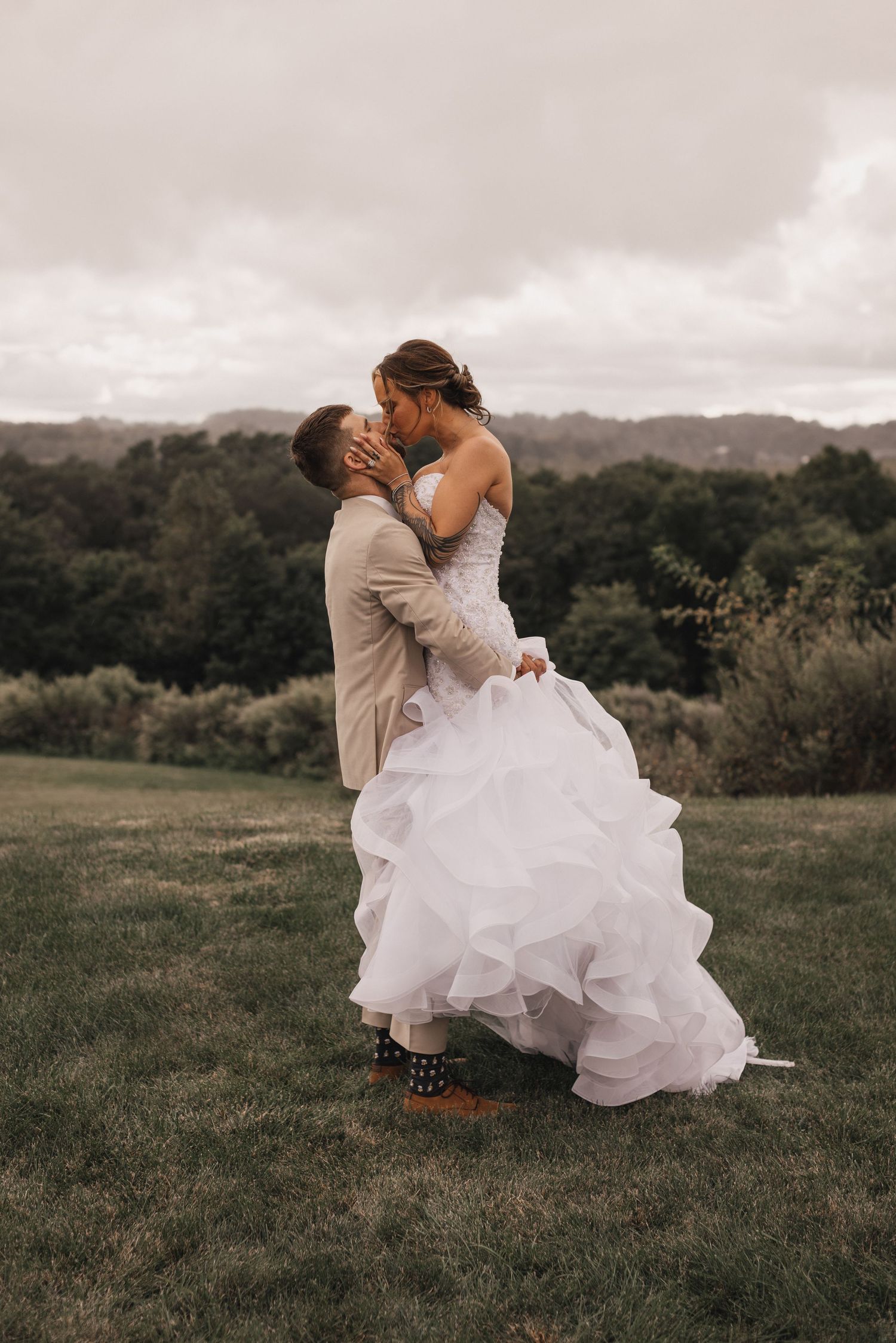 A bride in a flowing white dress crouches down emotionally in a grassy field during a wedding photoshoot.