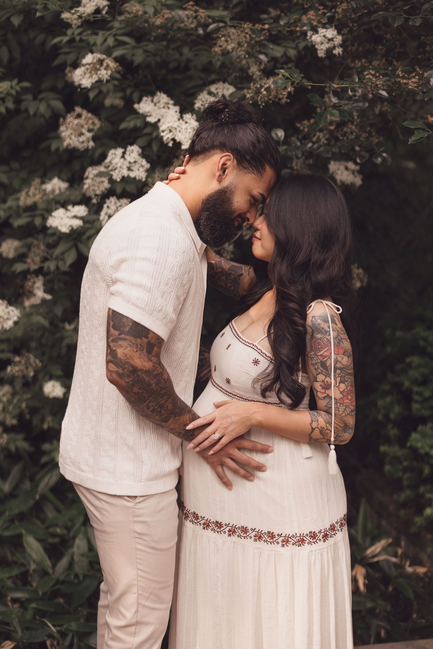 Maternity photos in white dress against flowering bushes in soft natural light.