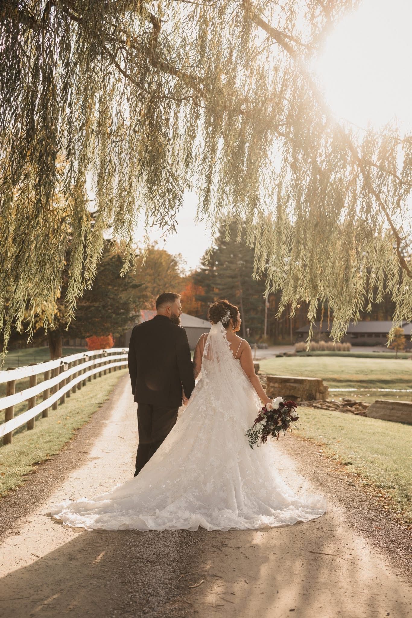 Romantic wedding photos under weeping willow trees along a white fence path at sunset.
