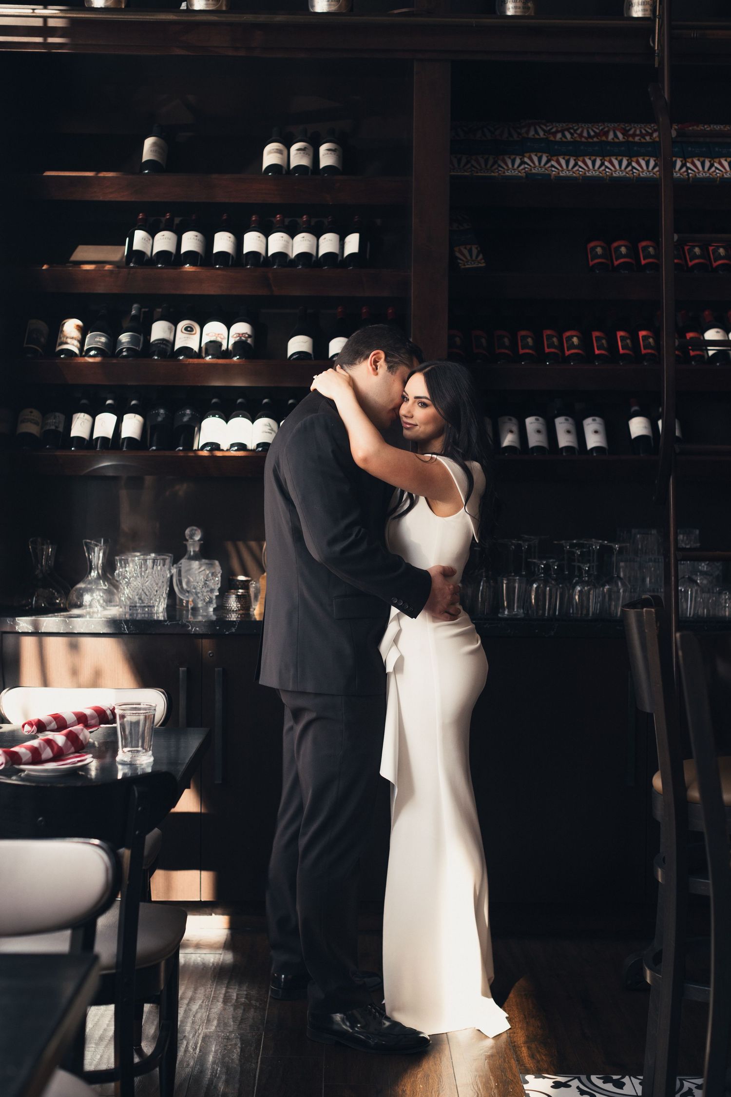 Romantic couples portrait series in a dimly lit wine cellar with bottle displays in background.