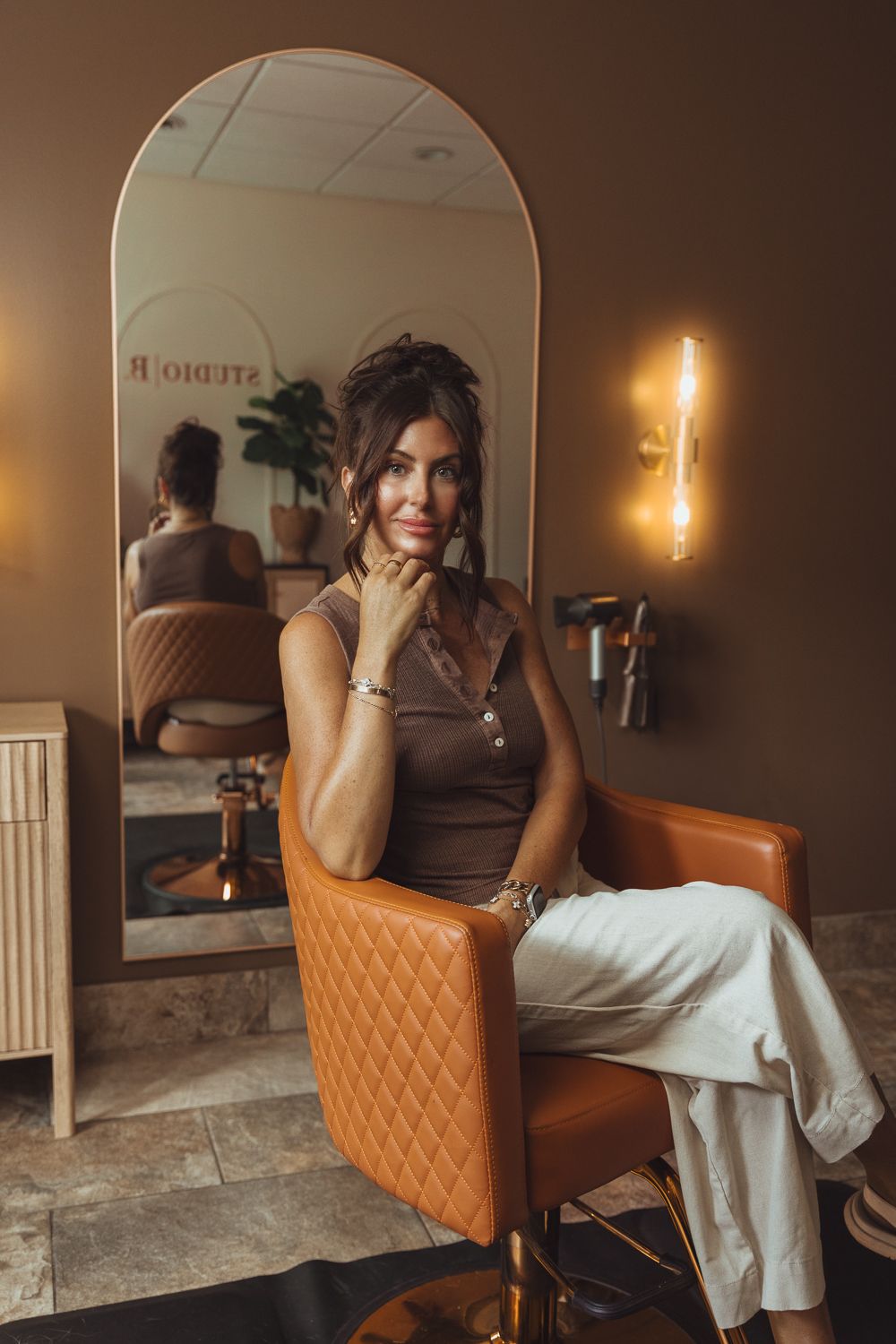 Woman sitting in orange leather salon chair poses thoughtfully in front of arched mirror at Studio R salon.