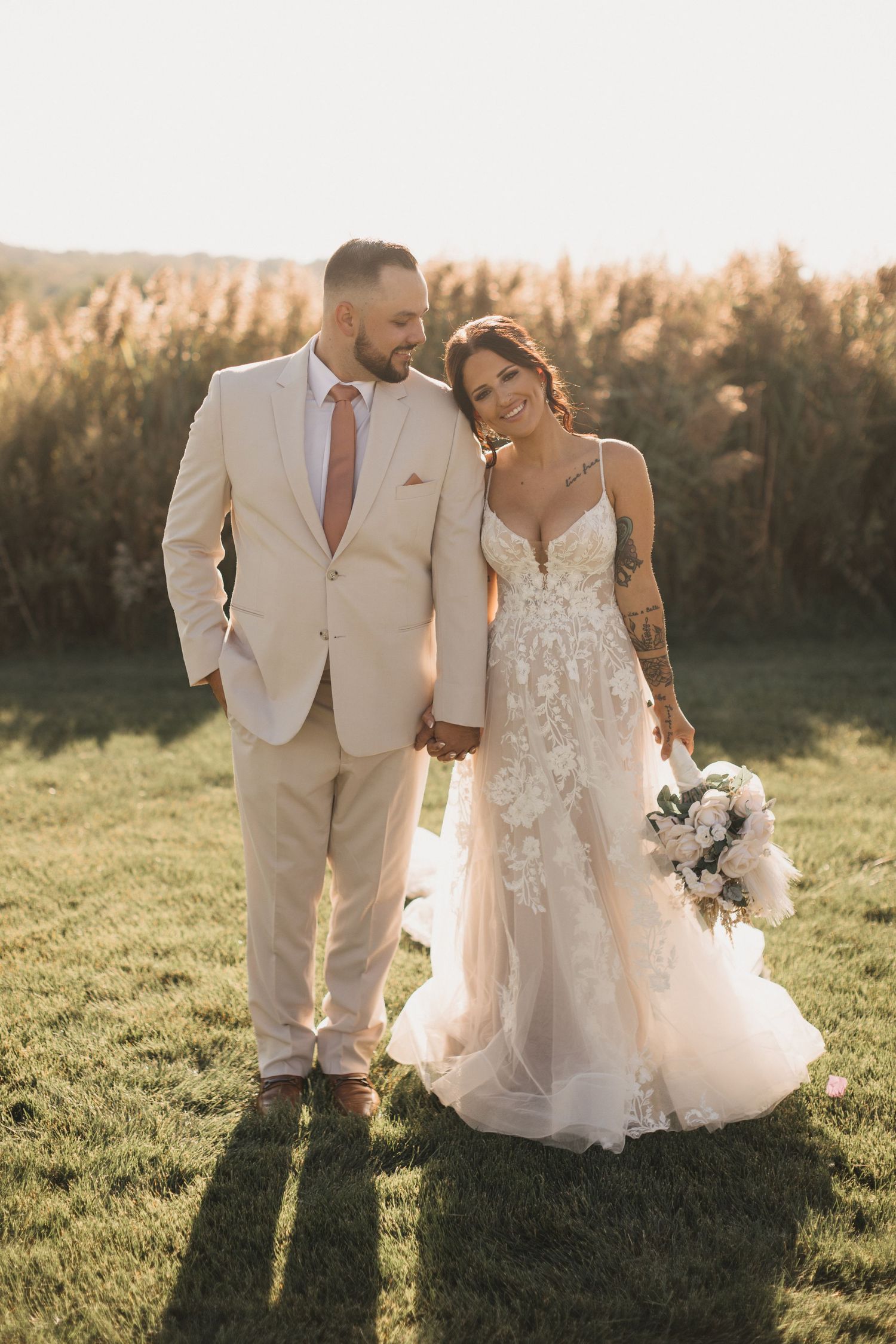 Newlyweds share intimate moments in a sunlit field wearing light-colored wedding attire.