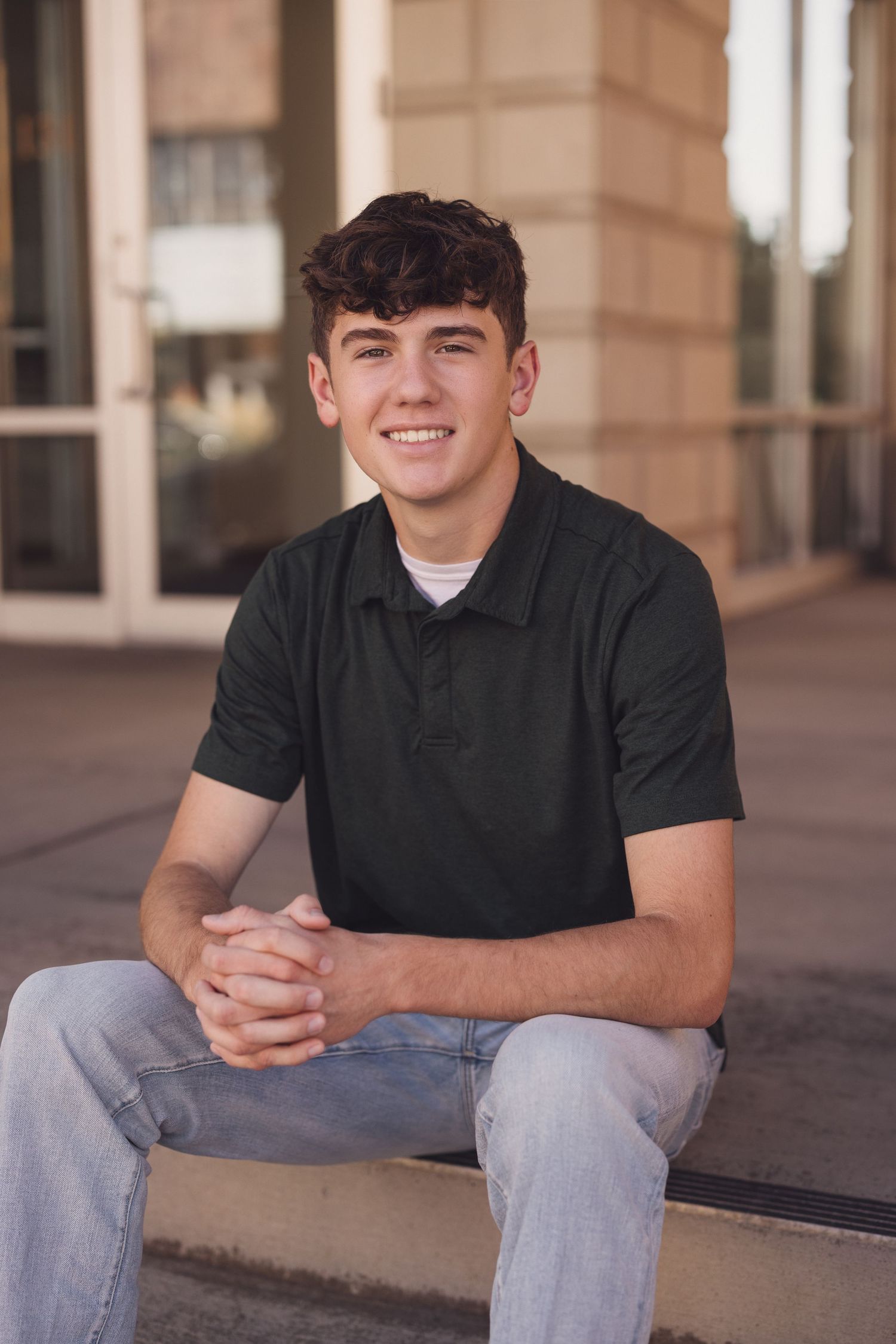 Young man in black polo and jeans sits casually on concrete steps near building columns.