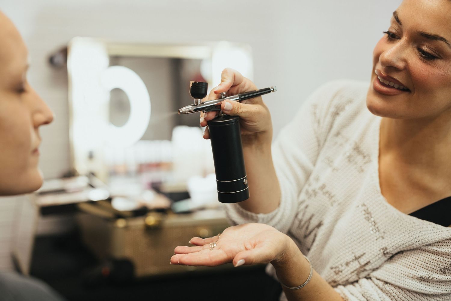 Makeup artist demonstrating airbrush tool to client in professional vanity mirror dressing room setting.